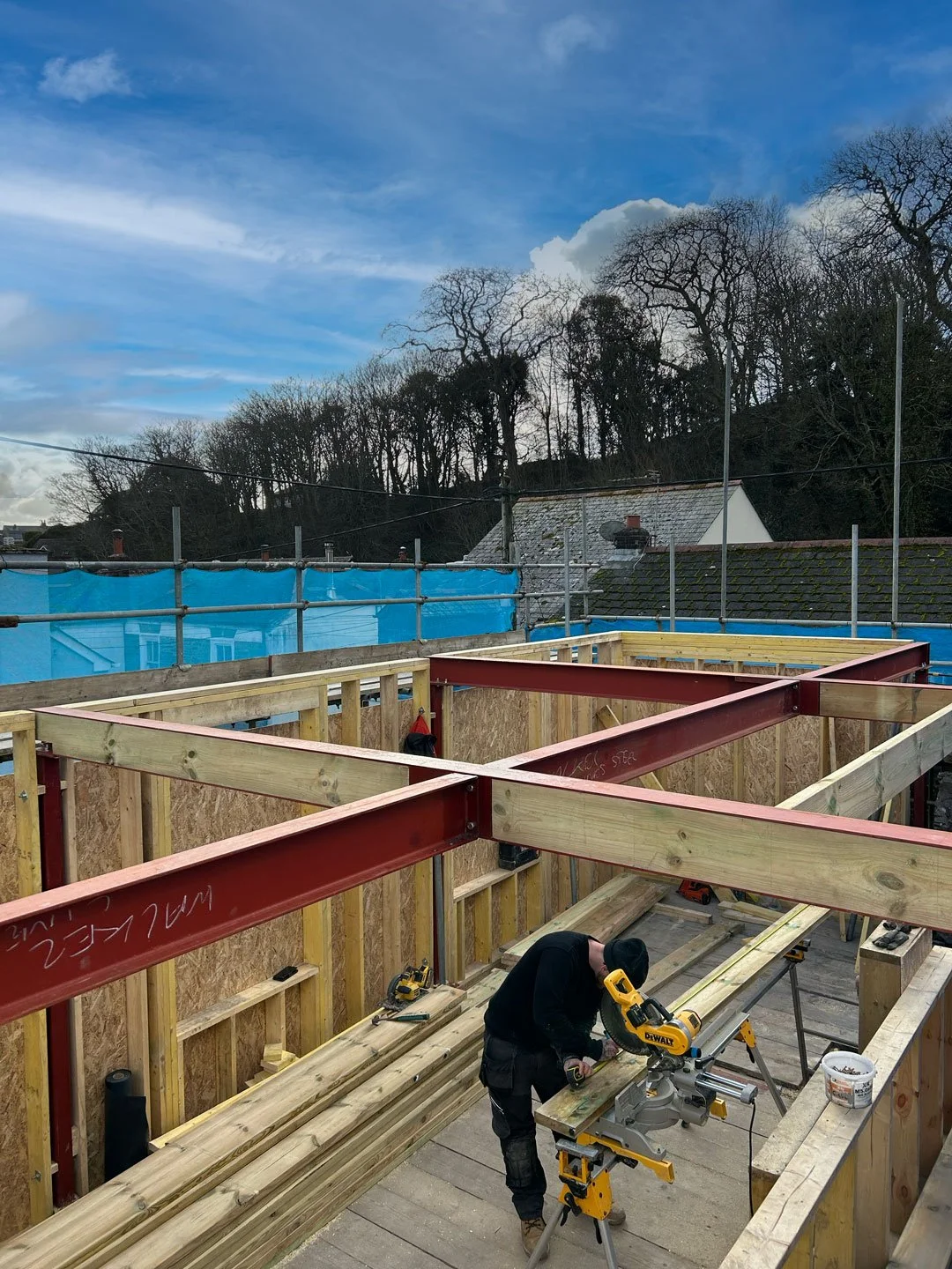 Construction site with wooden frame and steel beams, worker using a sliding miter saw, blue sky, trees and rooftops in the background.