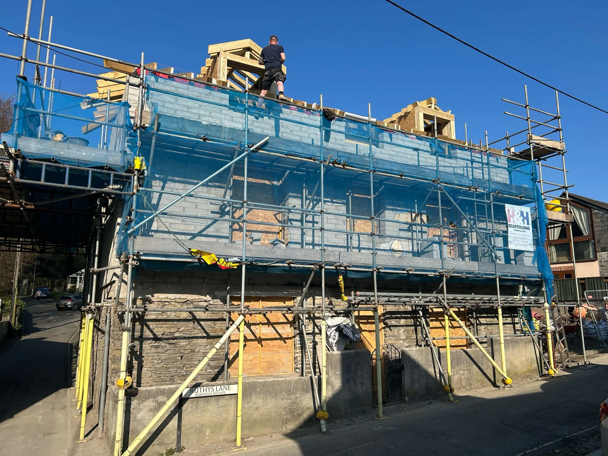 A house under construction with scaffolding around it, showing wooden framing on the roof and partially finished brick walls, against a bright blue sky.