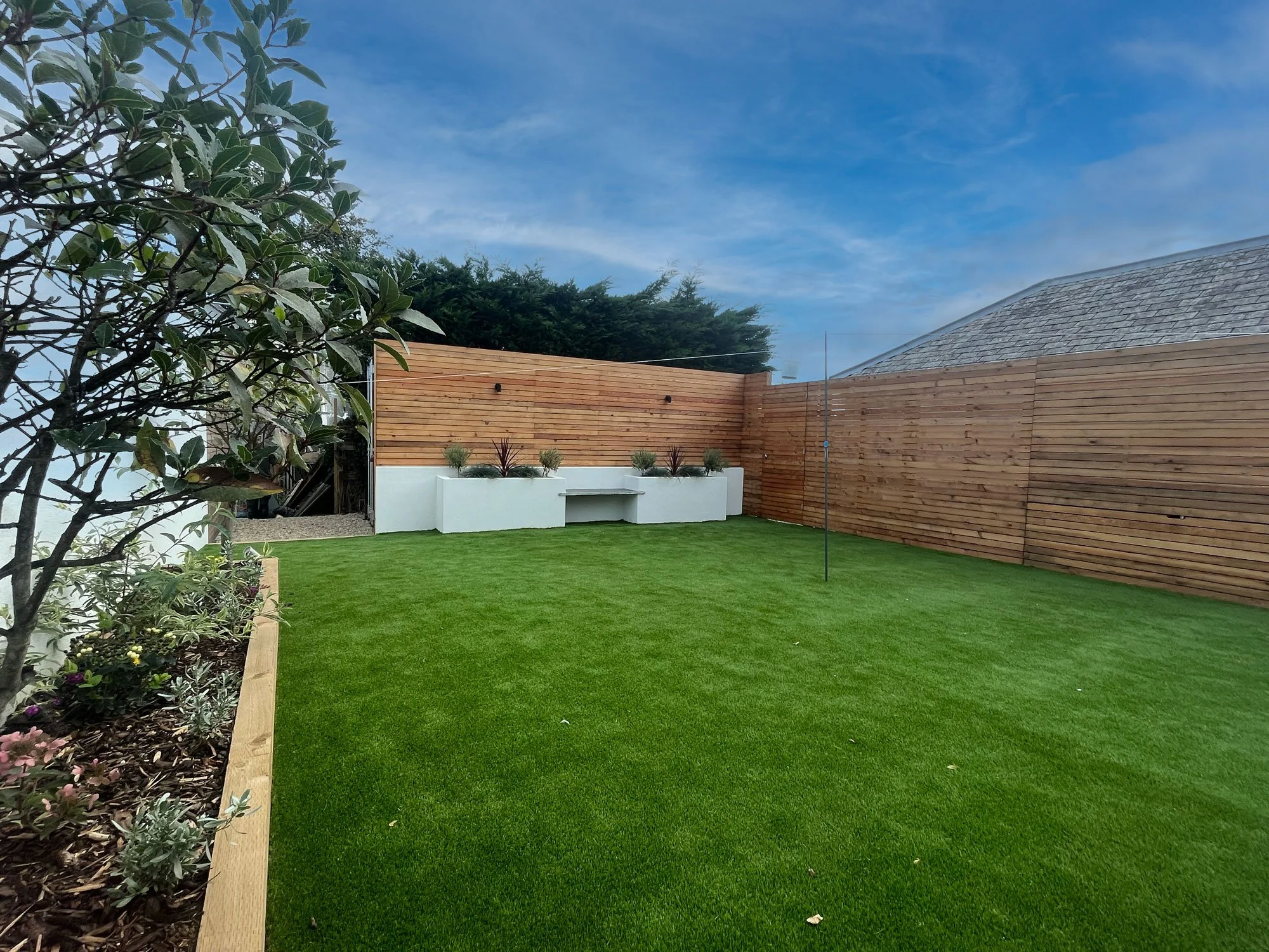 A backyard with a well-manicured green lawn, a wooden privacy fence, a white planter with succulents, and a corner planting bed with flowers and bushes. A sky with scattered clouds is visible above.