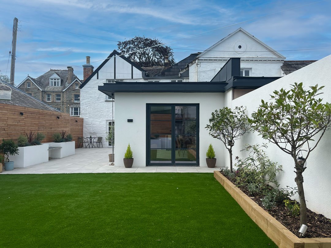 Modern backyard with a well-maintained lawn, potted plants, trees, and a small white building with black trim and glass doors, surrounded by a white wall and neighboring houses visible in the background.