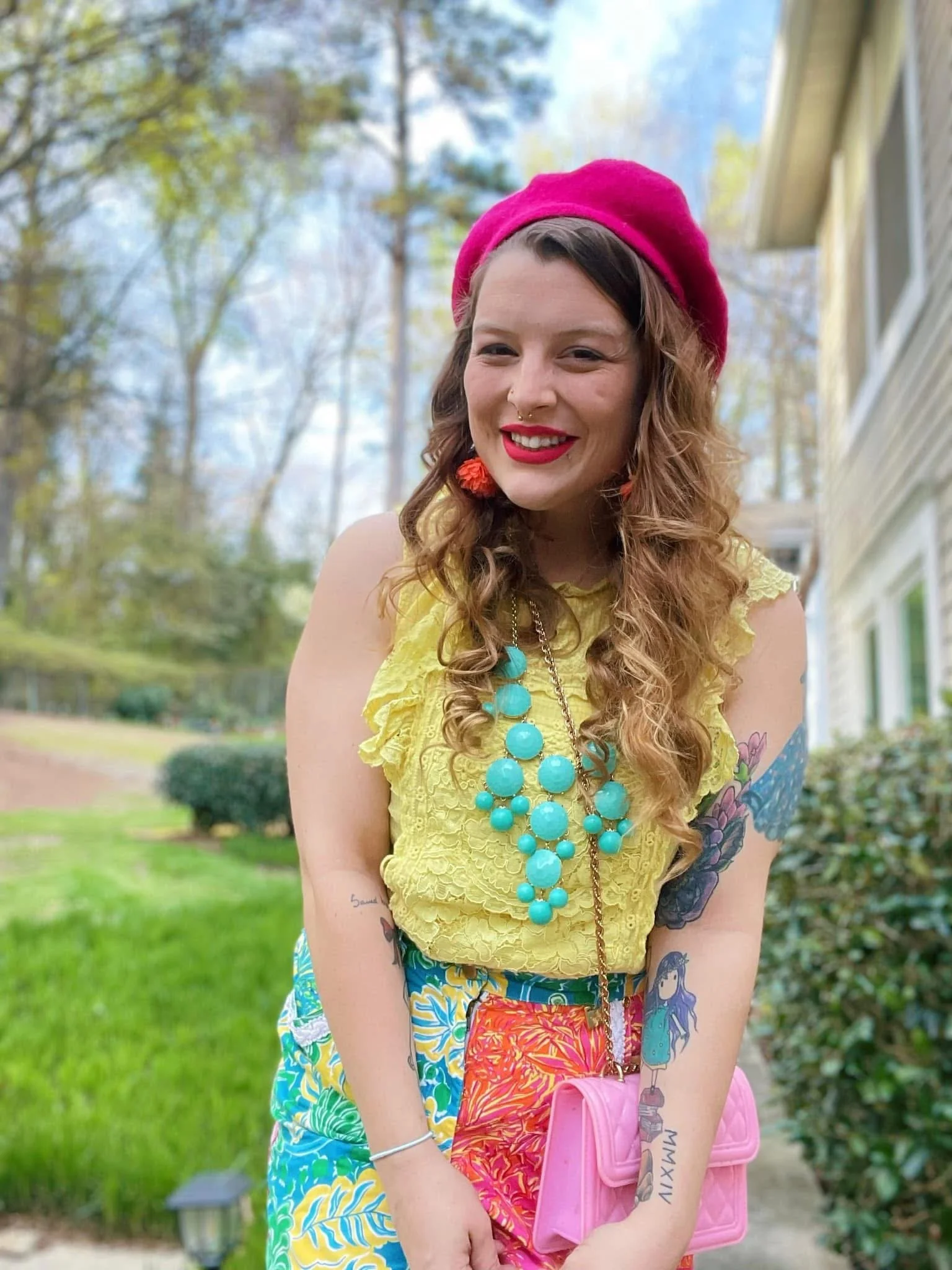 A smiling woman wearing a pink beret, yellow lace top, colorful patterned shorts, and turquoise jewelry, holding a pink purse outdoors with trees and a house in the background.