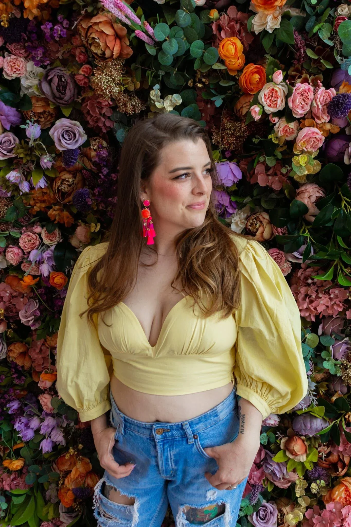 A woman with long brown hair and pink earrings standing in front of a colorful flower wall, wearing a yellow crop top and ripped blue jeans.
