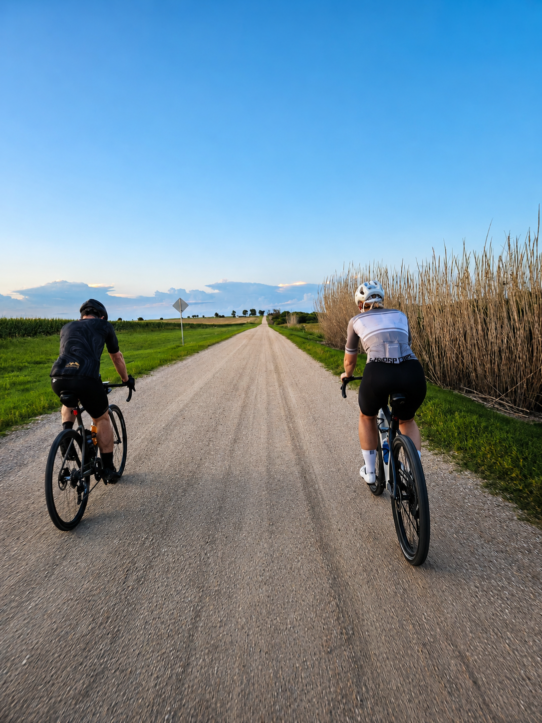 Two cyclists riding on a dirt road through a rural landscape with green fields, tall grass, and a blue sky with some clouds.