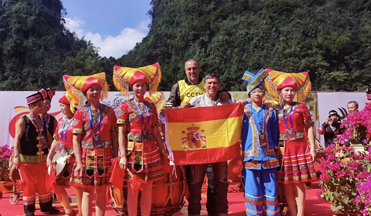 Grupo de personas con trajes tradicionales en un evento cultural, algunos sosteniendo una bandera de España, rodeados de flores y con un fondo de montañas.