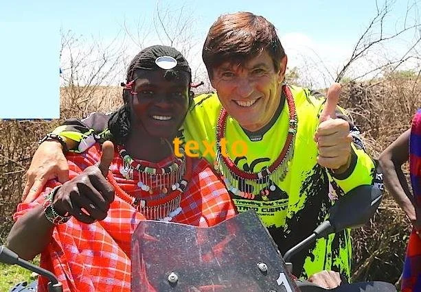 Dos personas sonrientes con ropa de color brillante posando juntas, una con gafas de sol y accesorios tradicionales, la otra con camiseta neon, en un ambiente al aire libre con vegetación seca.