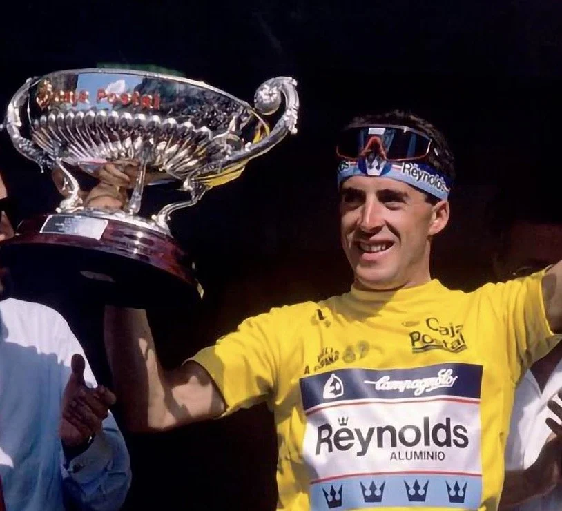 Un ciclista con camiseta amarilla sosteniendo un trofeo grande en forma de copa, celebrando un triunfo en una competencia de ciclismo.