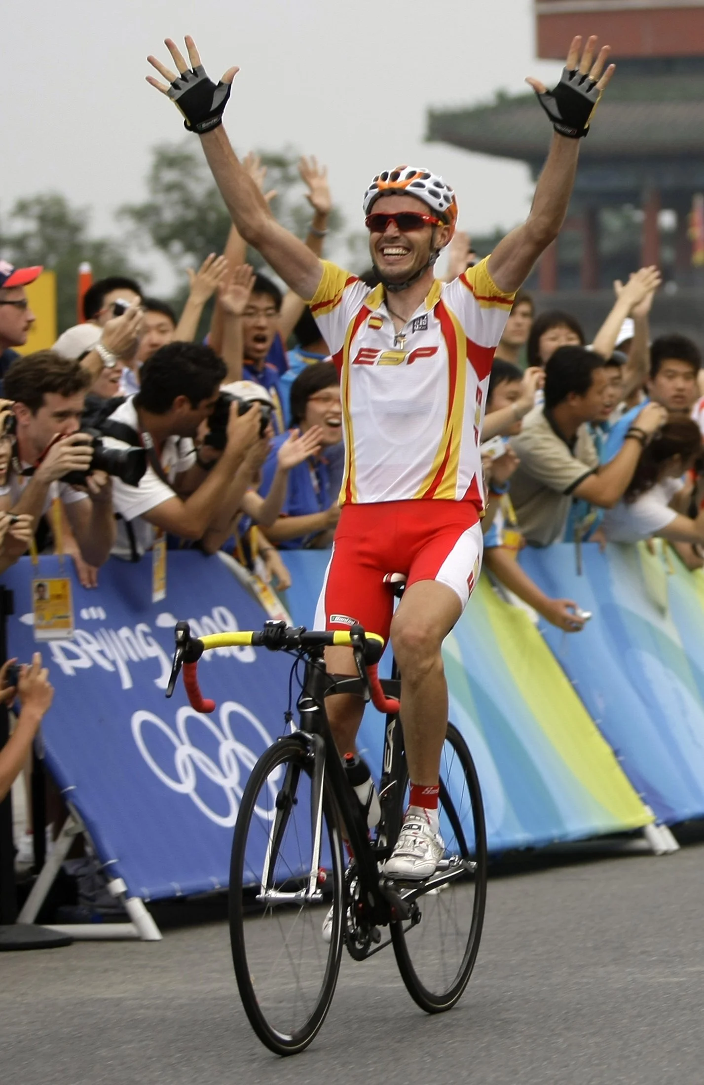 Un ciclista sonriendo con una camiseta blanca con detalles rojos y amarillos, y gafas de sol, levantando los brazos mientras pasa por un evento deportivo con gente celebrando y tomando fotos.