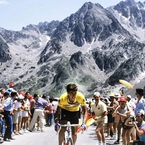 Un ciclista en una competencia en un camino montañoso rodeado de muchas personas, con montañas rocosas y picos nevados al fondo