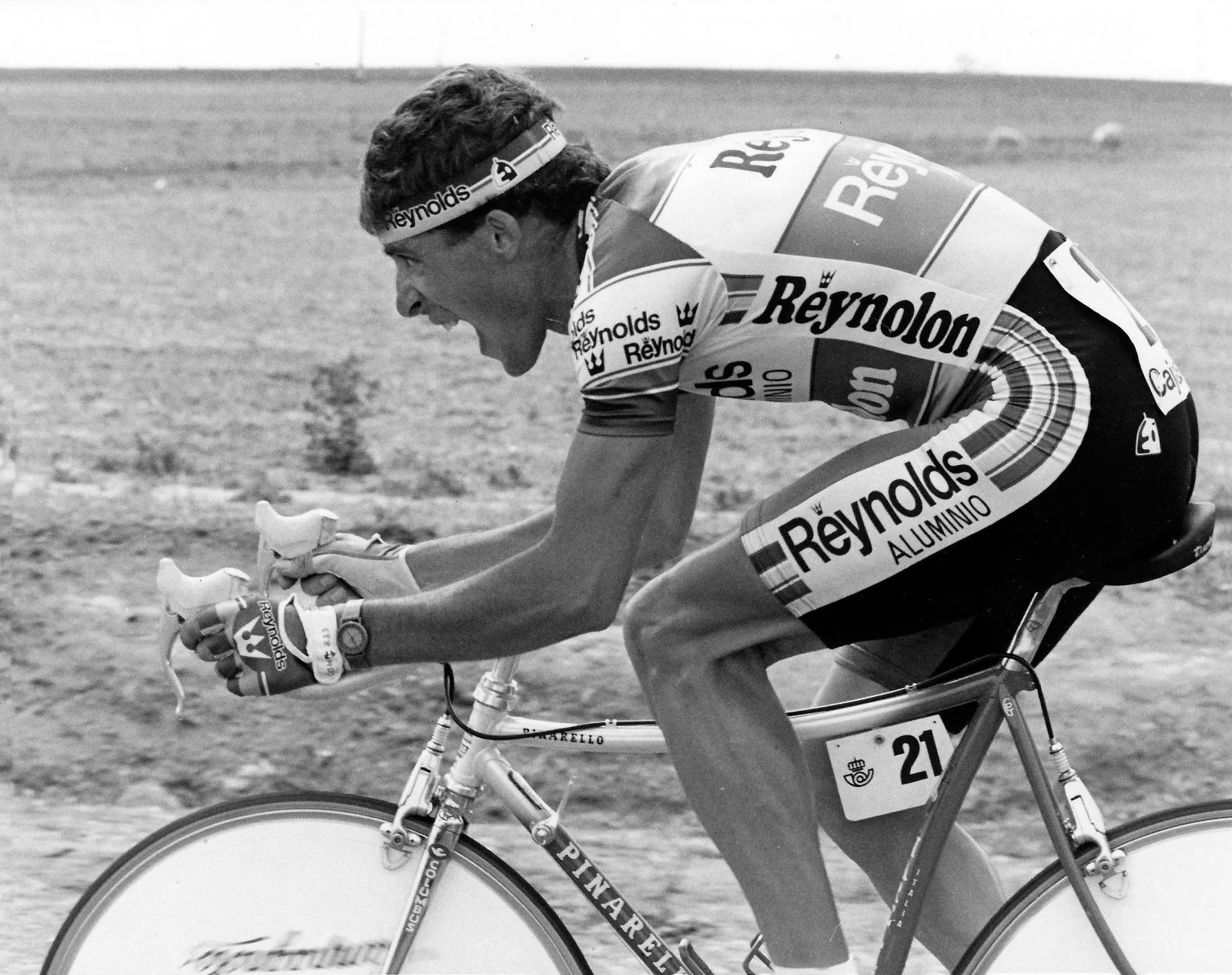 Ciclista en bicicleta durante una competición, con expresión de esfuerzo y concentración. Lleva uniforme con marcas de patrocinadores y guantes, en un fondo de tierra o césped.
