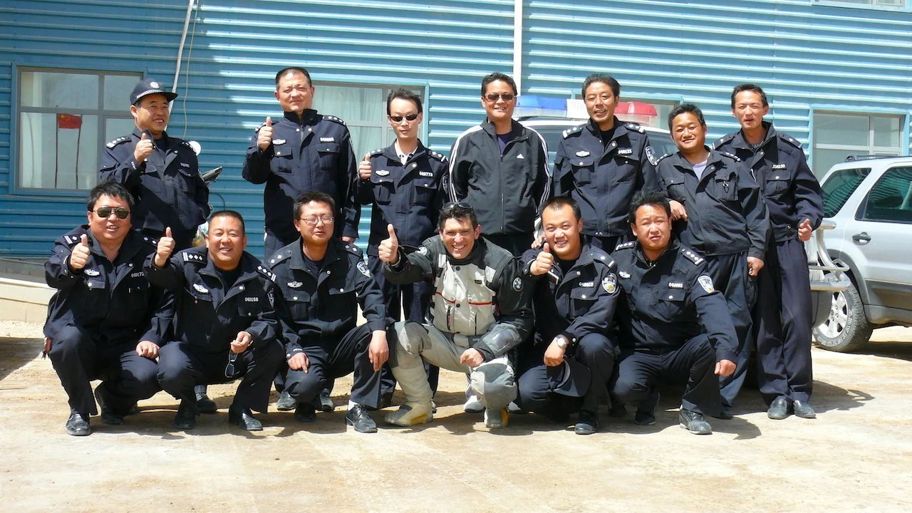 Grupo de policías en uniforme, posando y haciendo gestos de aprobación frente a un edificio de metal azul y un coche oficial en un día soleado.