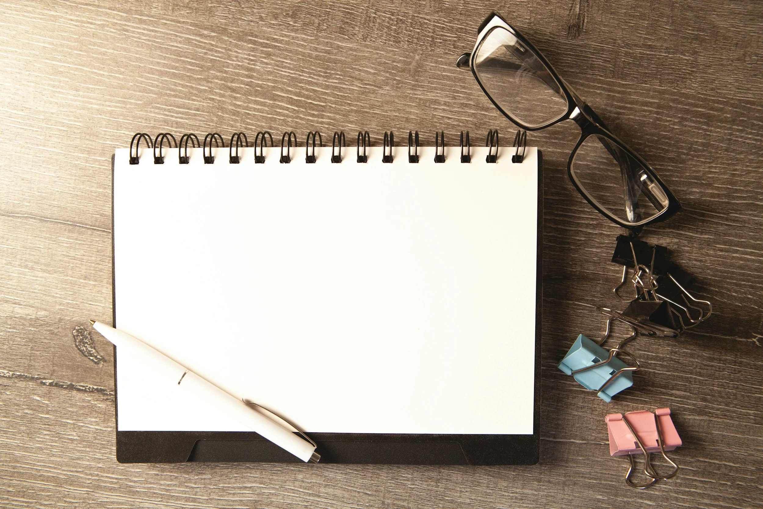 Desk with a closed laptop, a spiral notebook, glasses, paper clips, a pen, and sticky notes on a wooden surface.