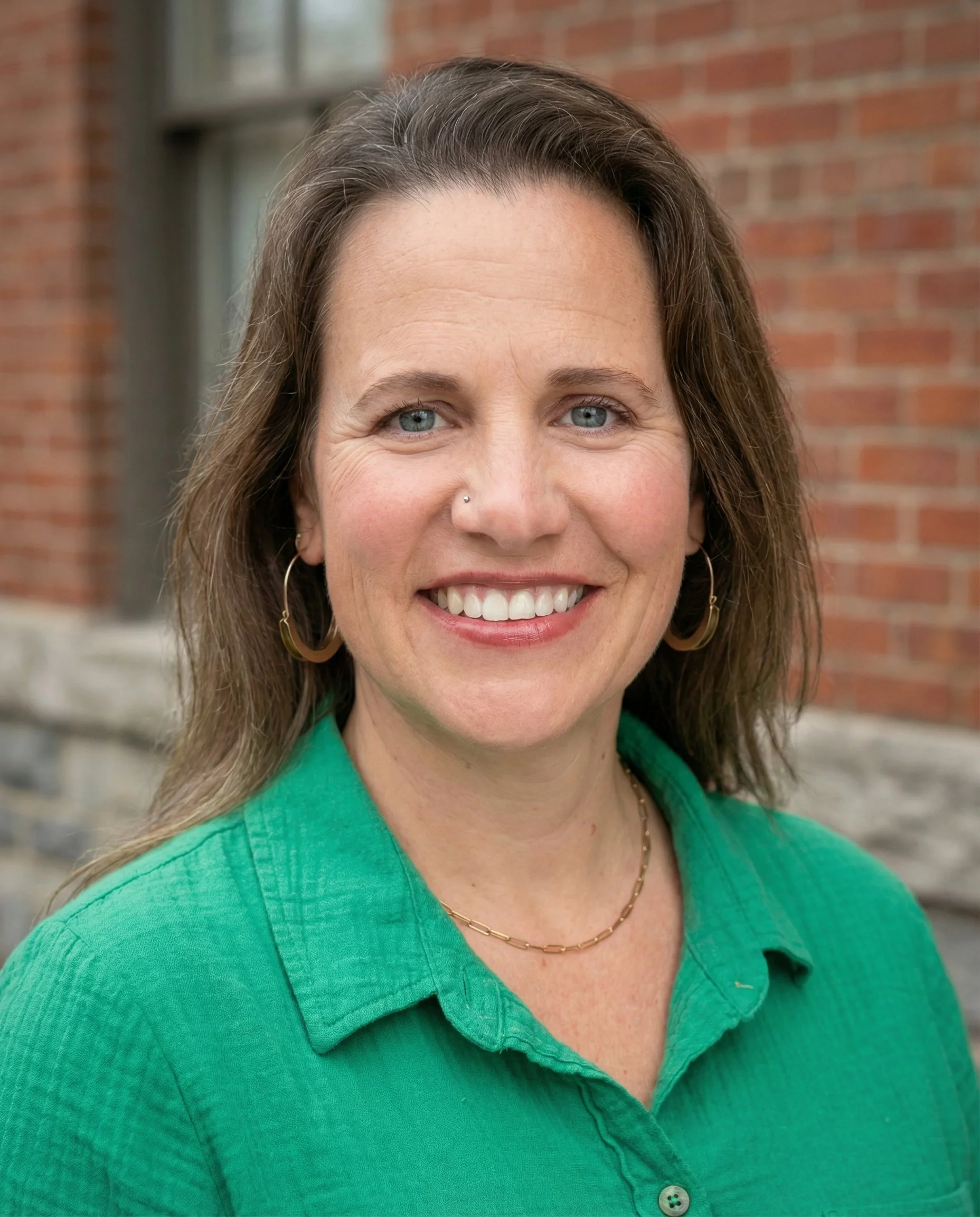 Close-up of a smiling woman with brown, wavy hair, blue eyes, wearing hoop earrings, a gold chain necklace, and a green button-up shirt, standing outdoors in front of a brick building.