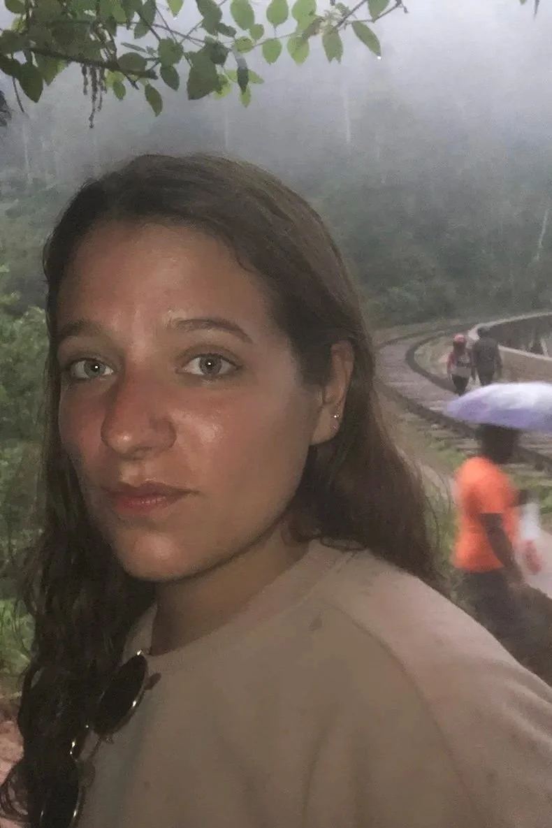 A woman with long brown hair and light-colored eyes taking a selfie outdoors on a rainy day, with people walking in the background and holding umbrellas.