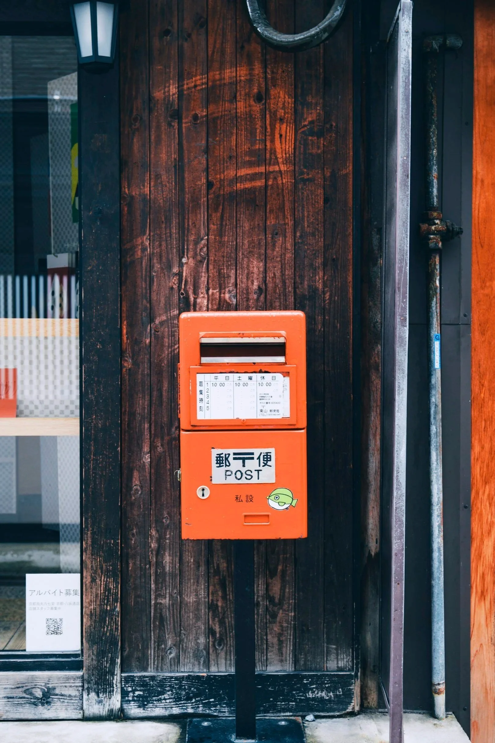 Orange mailbox mounted on a wooden wall with a sticker of a green fish character and Japanese writing.
