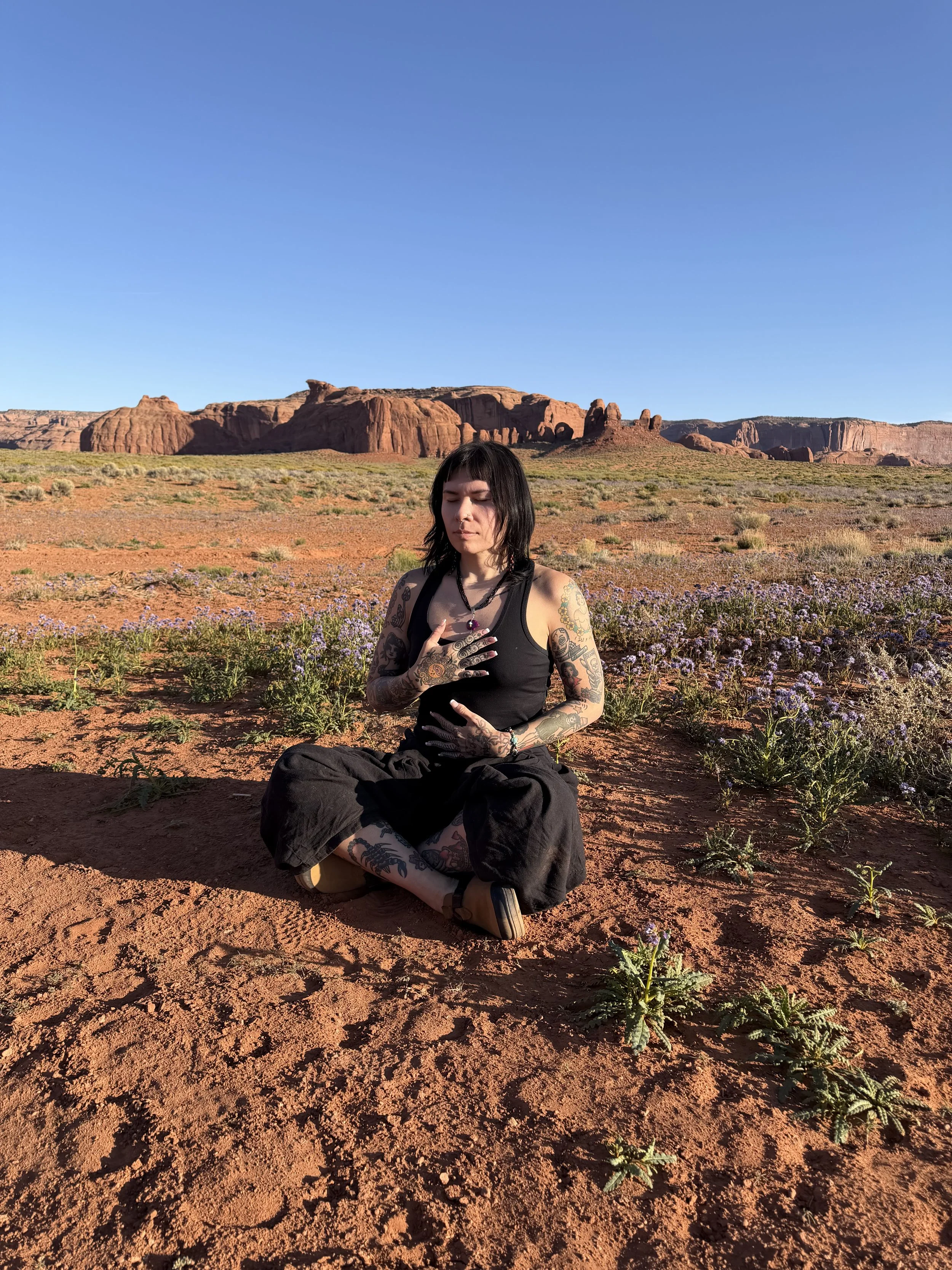 A woman with tattoos, black hair, and tattoos on her arms and legs, sitting cross-legged in a desert landscape with red rocks and purple flowers, under a clear blue sky.
