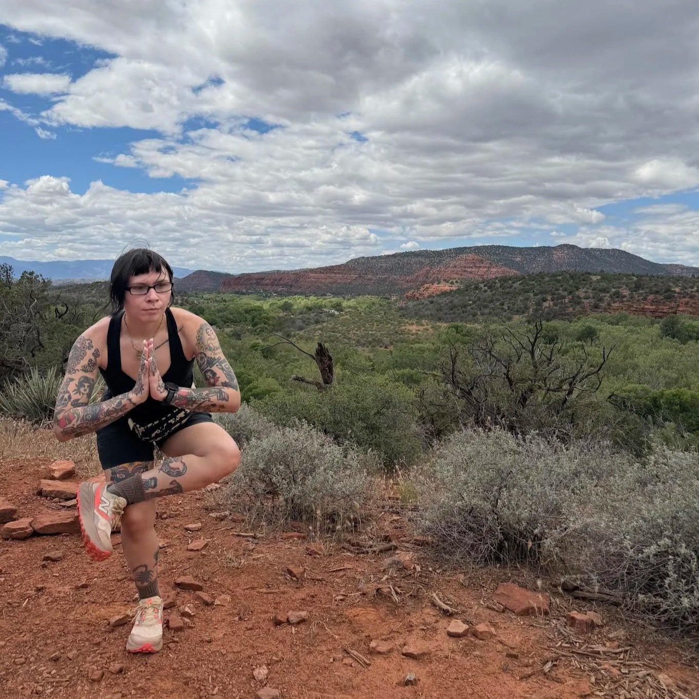 A woman with tattoos wearing glasses in half chair pose outdoors on a dirt trail with a scenic landscape of green bushes, trees, red rock formations, and a cloudy sky in the background.