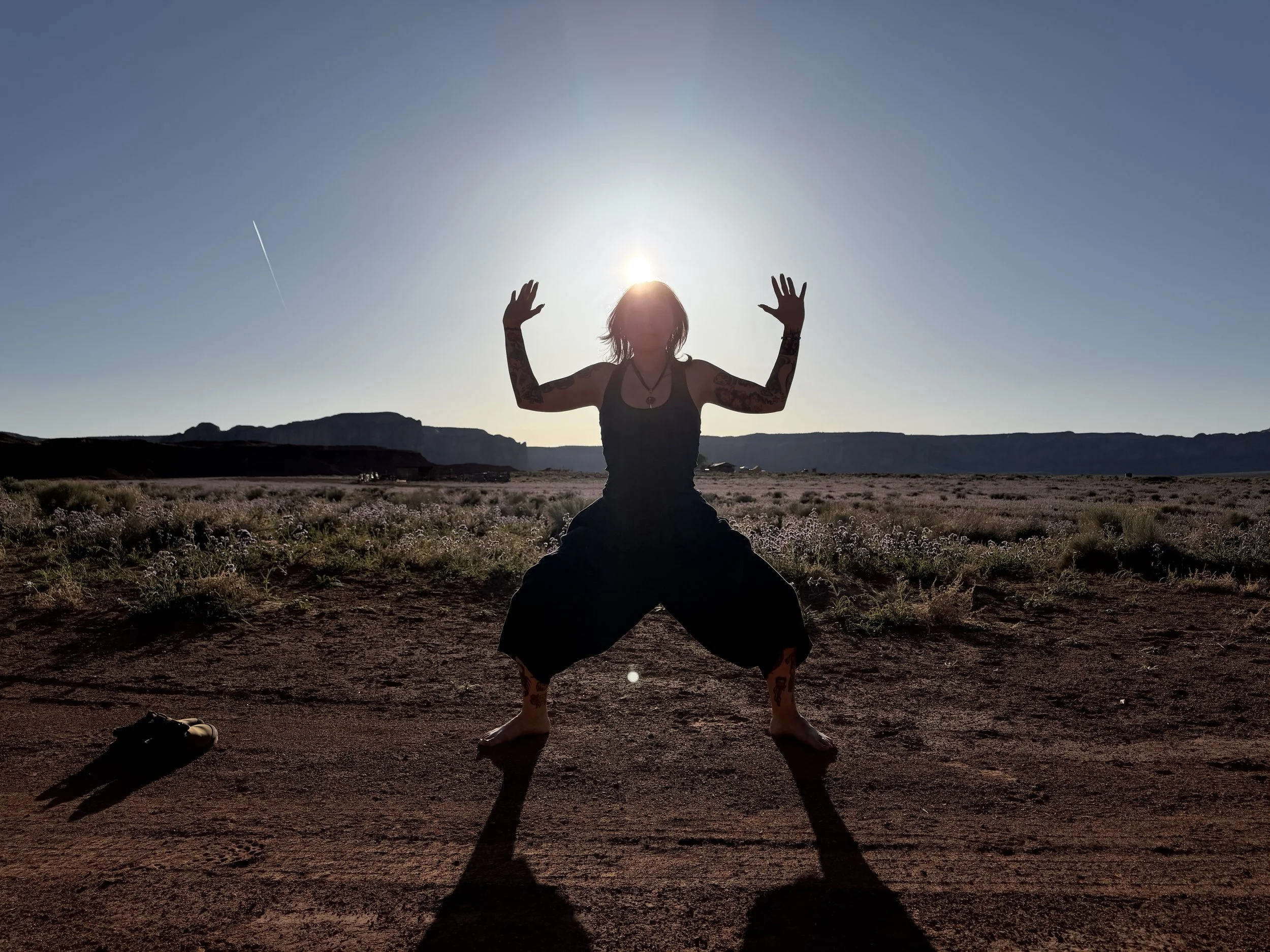 A woman practicing yoga outdoors during sunset in a desert landscape, in Goddess pose, casting a long shadow.