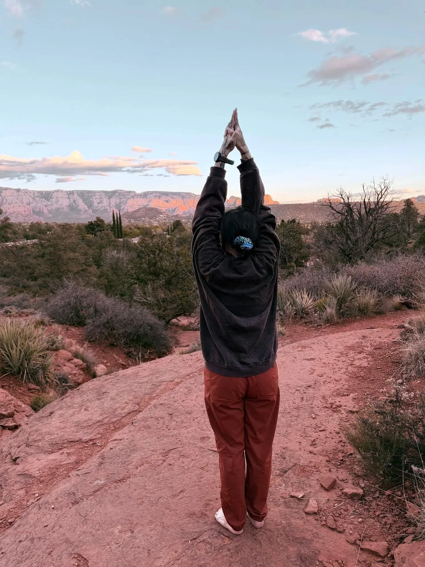 Person standing outdoors on a rocky trail with arms raised, hands together in Kali mudra, overlooking a desert landscape with trees and mountains, under a sky with clouds.