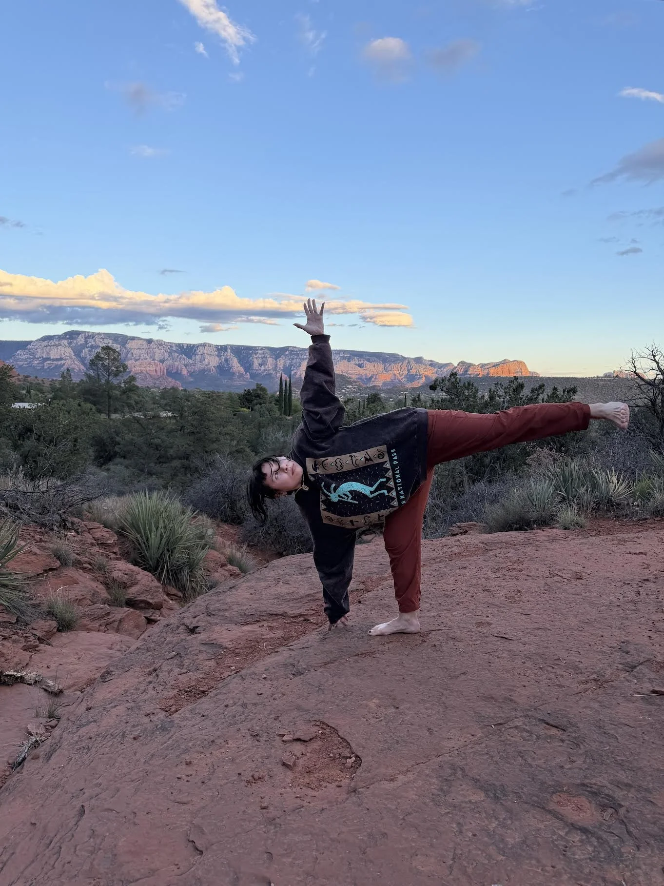 Person practicing half moon pose outdoors on a rocky terrain, with a desert landscape and mountains in the background, under a partly cloudy sky.