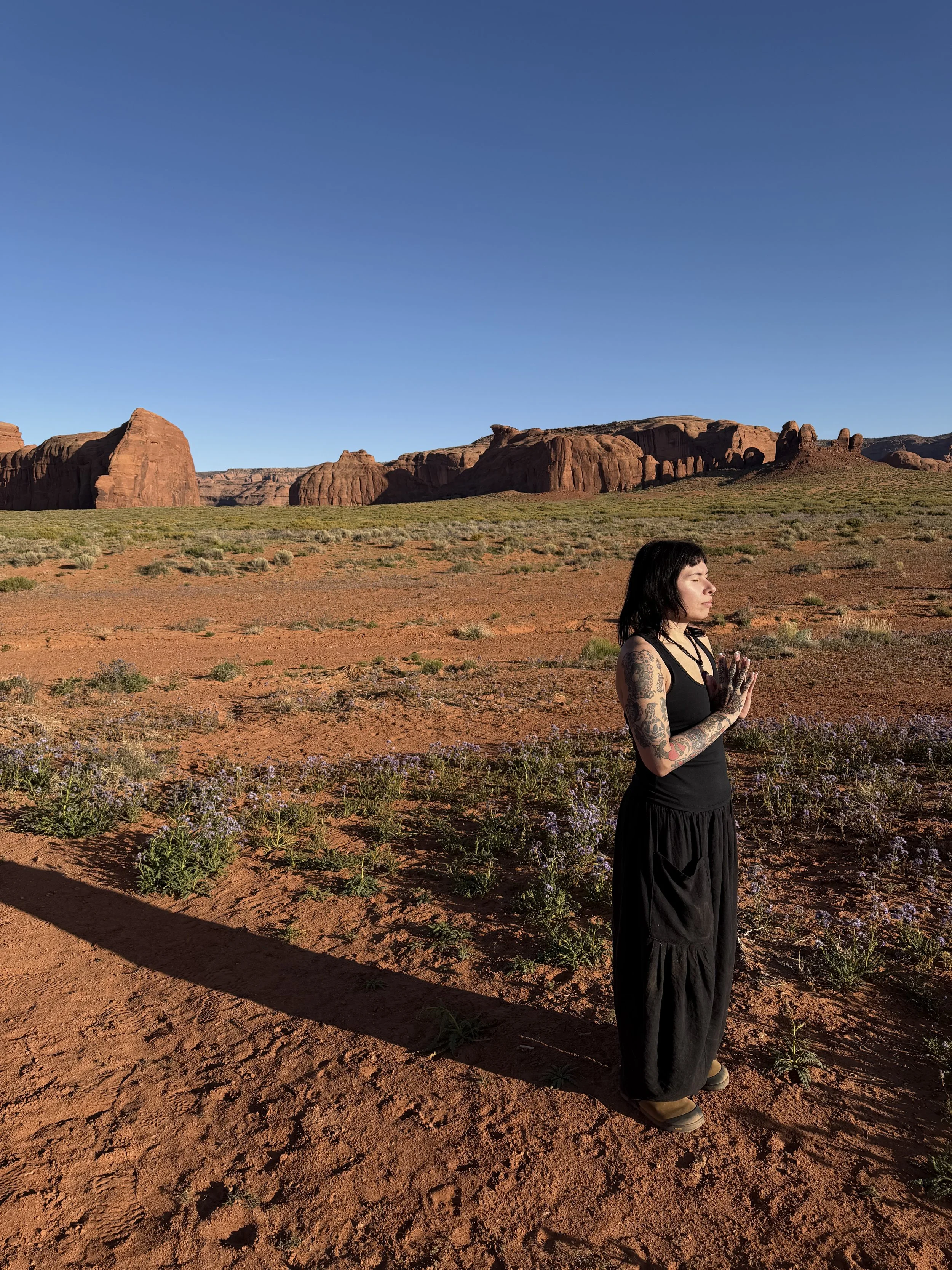 A woman with tattoos on her arms and black hair, wearing a black sleeveless top and long black skirt, practicing meditation with her eyes closed and hands in a prayer position, standing on reddish desert soil with sparse vegetation, under a clear blue sky, with red rock formations in the background.