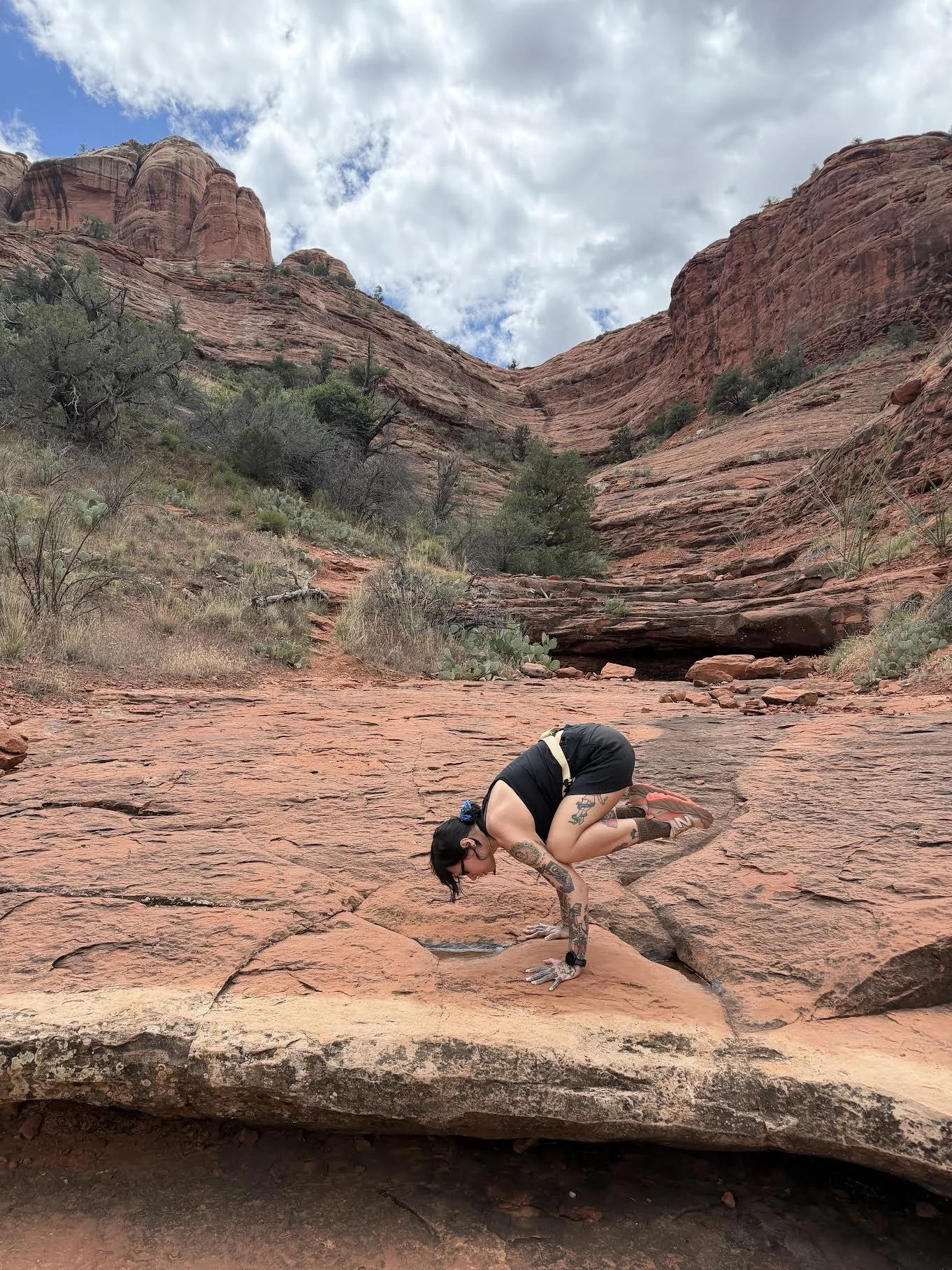 A woman with tattoos practicing crow pose on a red rock surface in a desert landscape with large red rock formations and a partly cloudy sky overhead.