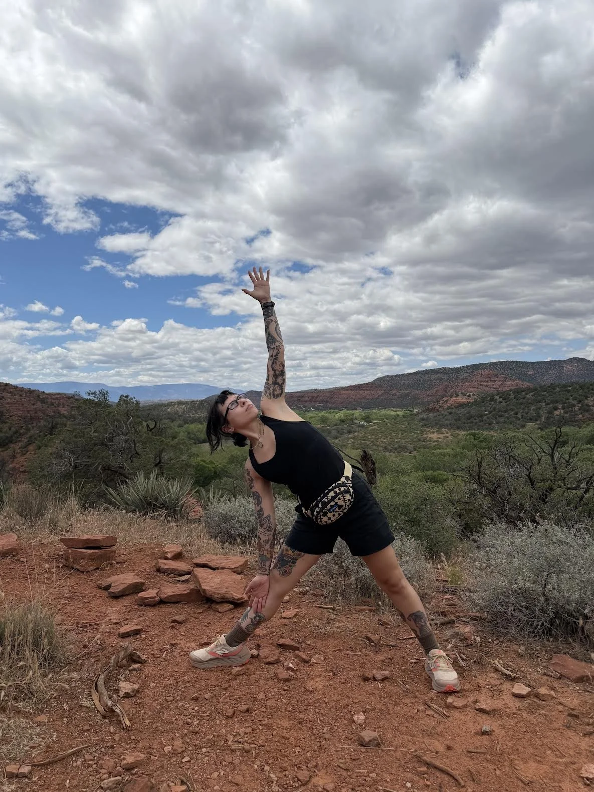 A woman with tattoos and glasses practicing triangle pose outdoors on a rocky, reddish trail with a scenic view of green trees, hills, and a cloudy sky in the background.