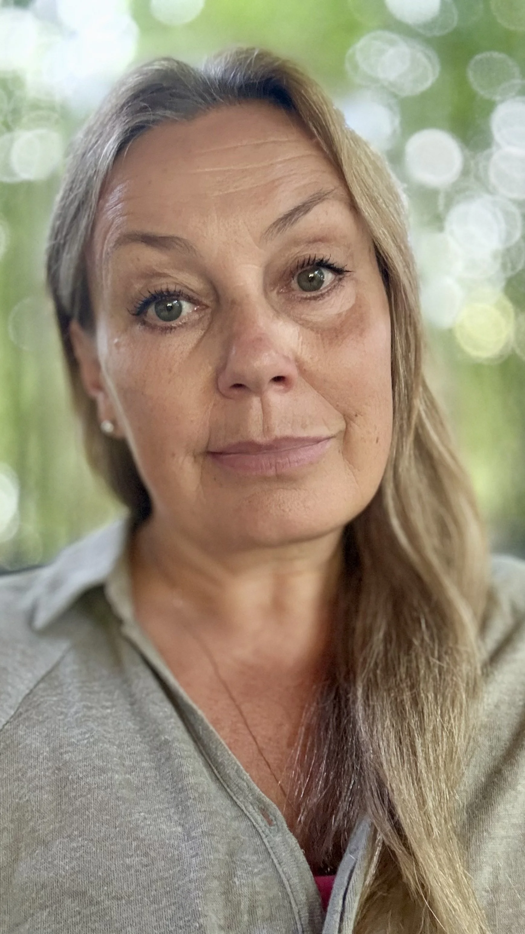 A woman with long, light brown hair and hazel eyes, wearing a light gray zip-up hoodie, standing outdoors with blurred green foliage in the background.