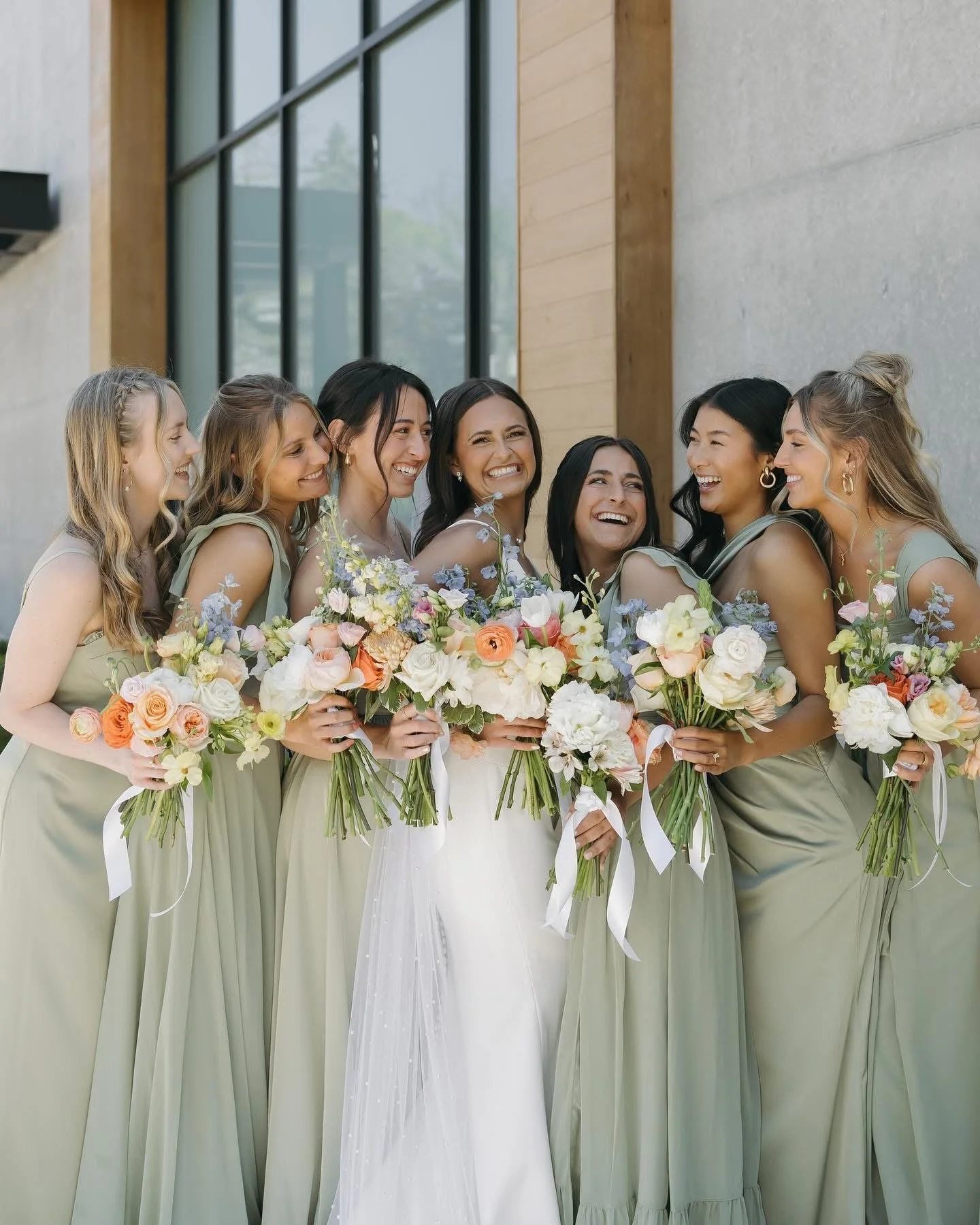 A group of eight women dressed in matching light green dresses, standing together outside, holding bouquets of colorful flowers, smiling and laughing.