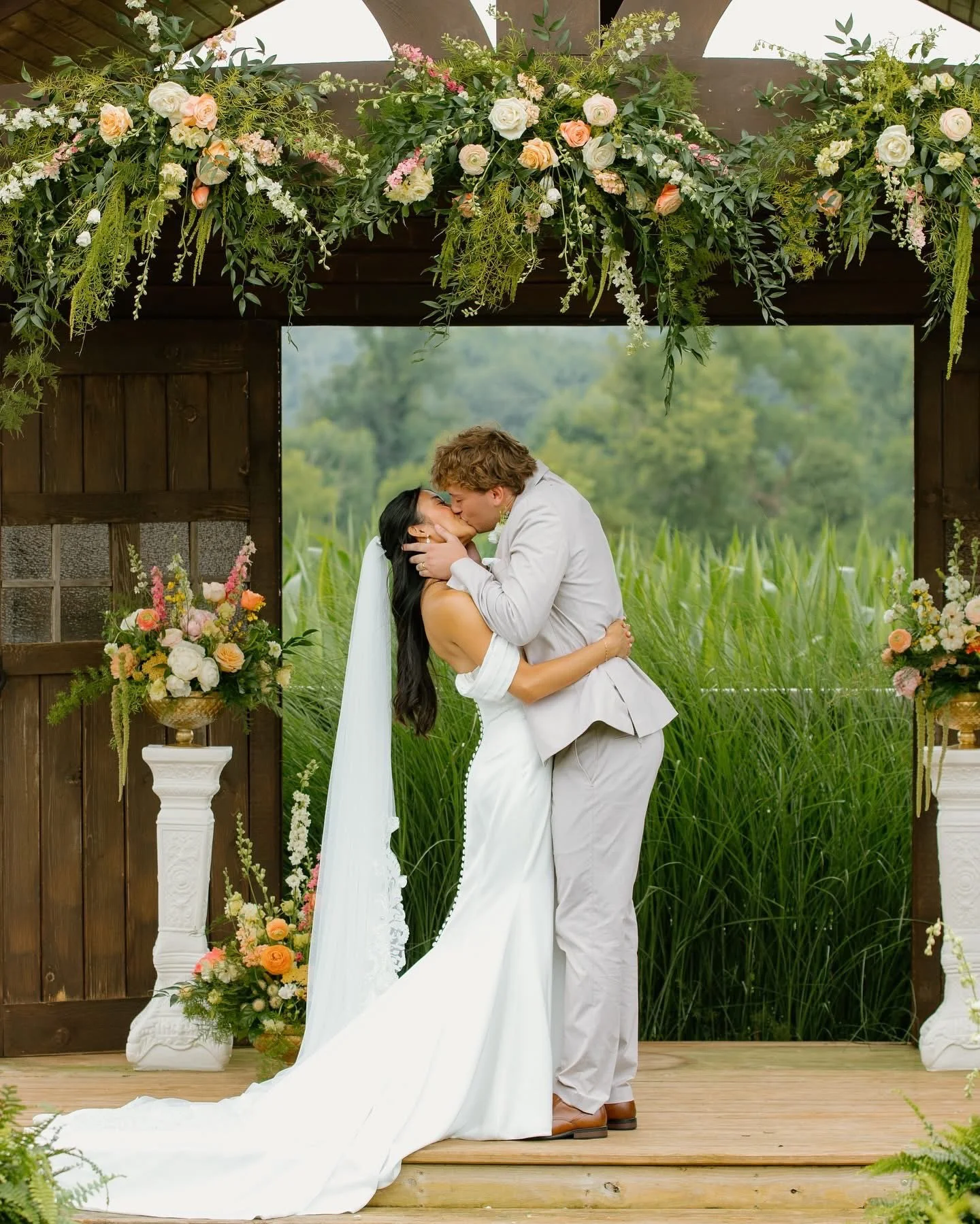 A bride and groom kiss during their outdoor wedding ceremony, standing on a wooden platform surrounded by greenery and floral arrangements.