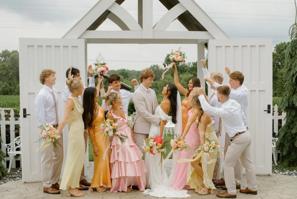 A wedding celebration with a bride and groom standing in front of a white gate, surrounded by bridesmaids and groomsmen, some raising their fists, outdoors with greenery and cloudy sky in the background.
