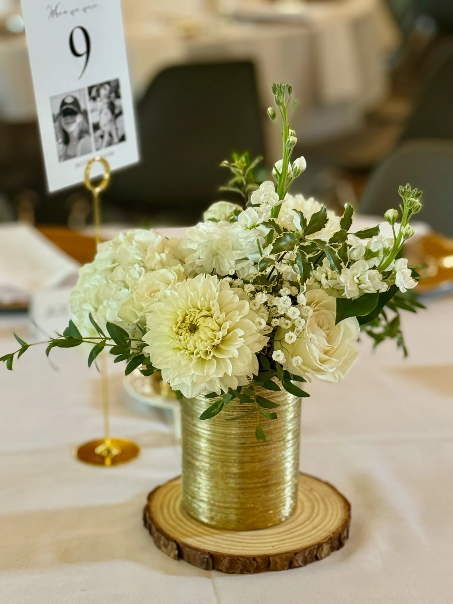 A floral centerpiece with white flowers and greenery in a gold textured vase, placed on a wood slab.
