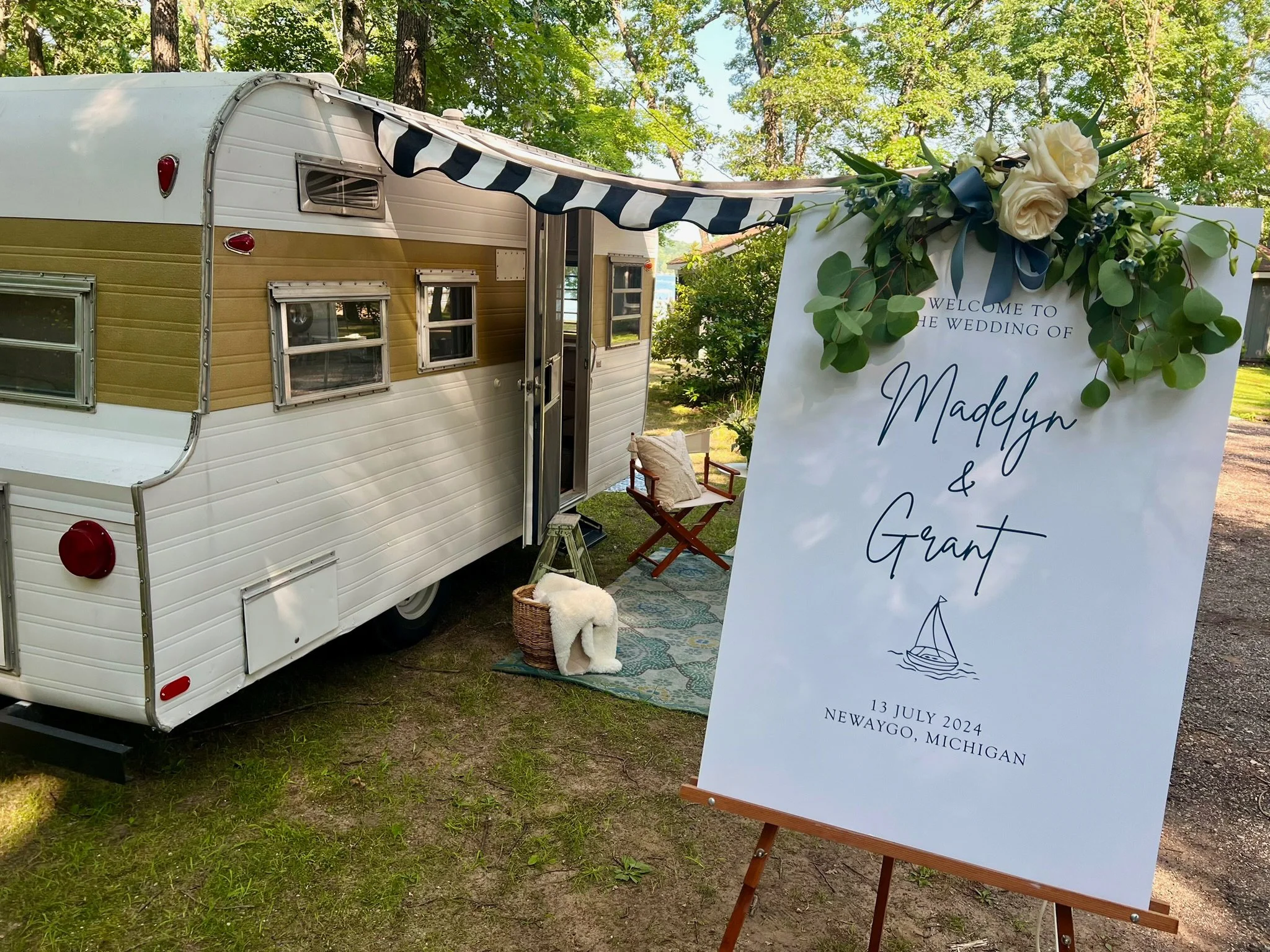 A vintage camper trailer decorated for a wedding with a floral arrangement on a large welcome sign. The sign reads 'Welcome to the wedding of Madelyn & Grant' and includes the date, location, and an illustration of a sailboat. The scene is outdoors with trees and greenery in the background.