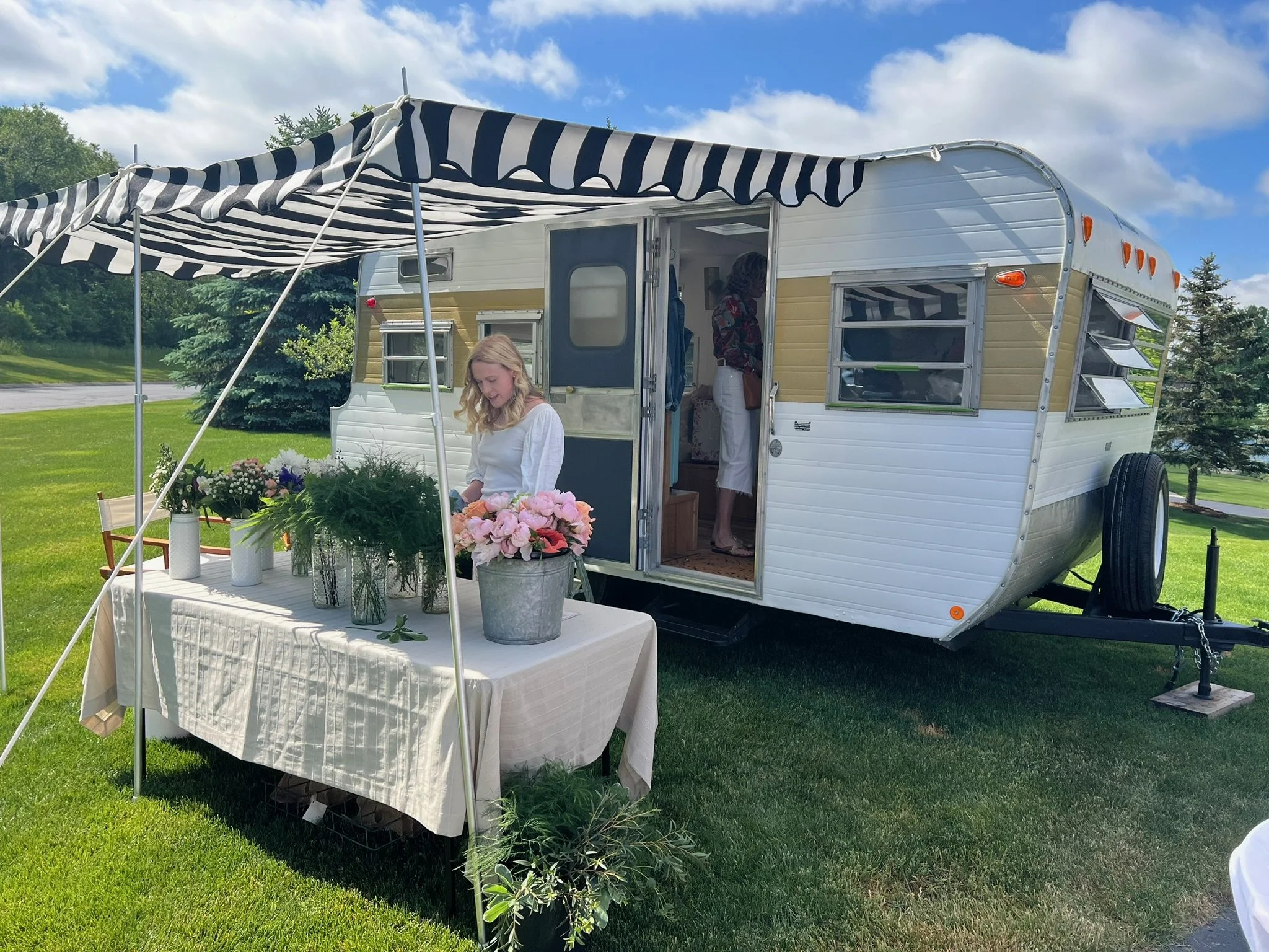 A vintage camper trailer set up outdoors with a black and white striped canopy. There is a table with flowers in vases, and a woman is sitting at the table. Inside the camper, two people are visible, one standing and one sitting. The scene is on a gr