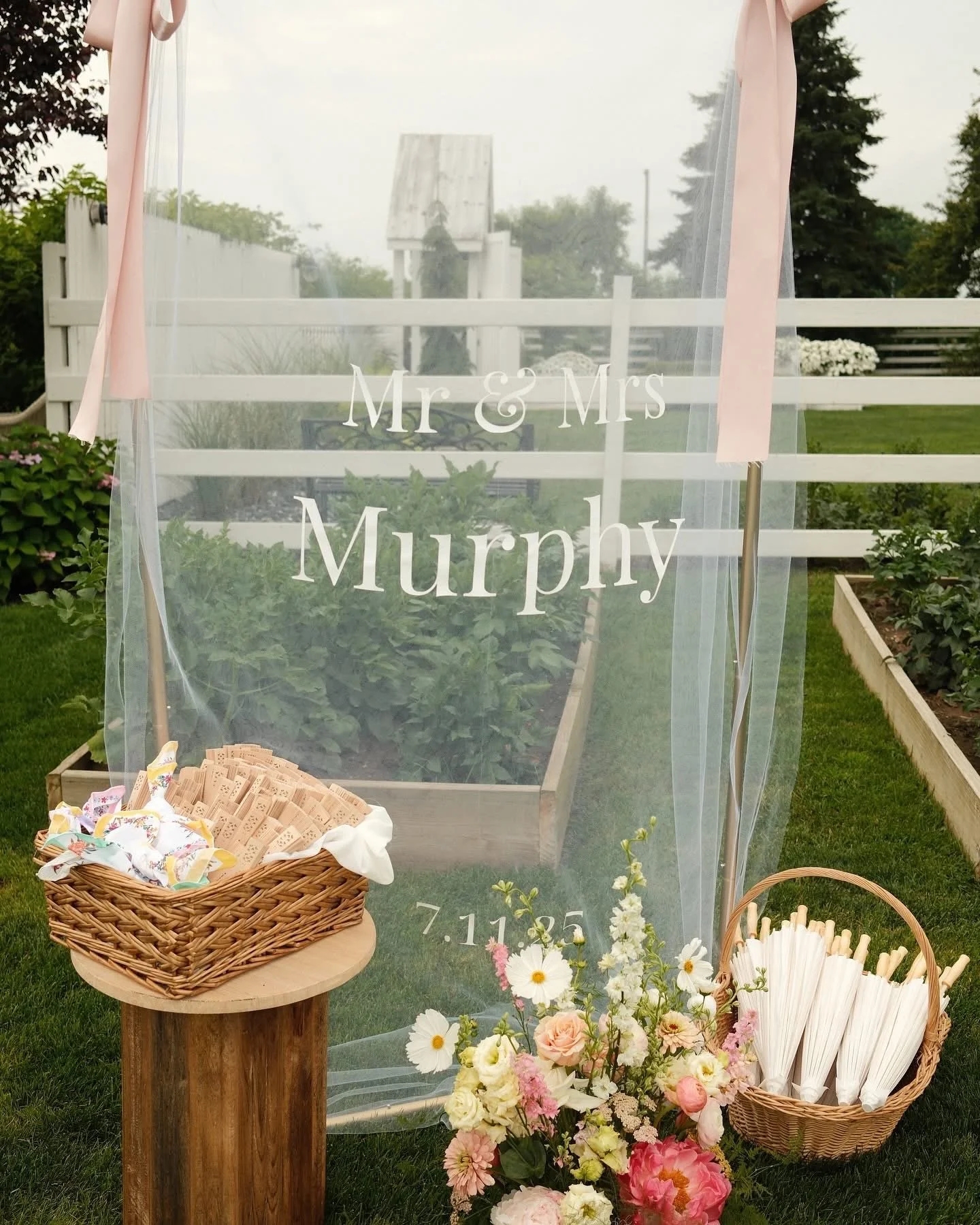 A wedding setup outdoors with a transparent veil that has 'Mr & Mrs Murphy' and the date '7.11.85' written on it. There is a small wooden table with a basket of pre-cut items, surrounded by flowers and a basket of cocktail stirrers or straws. The bac