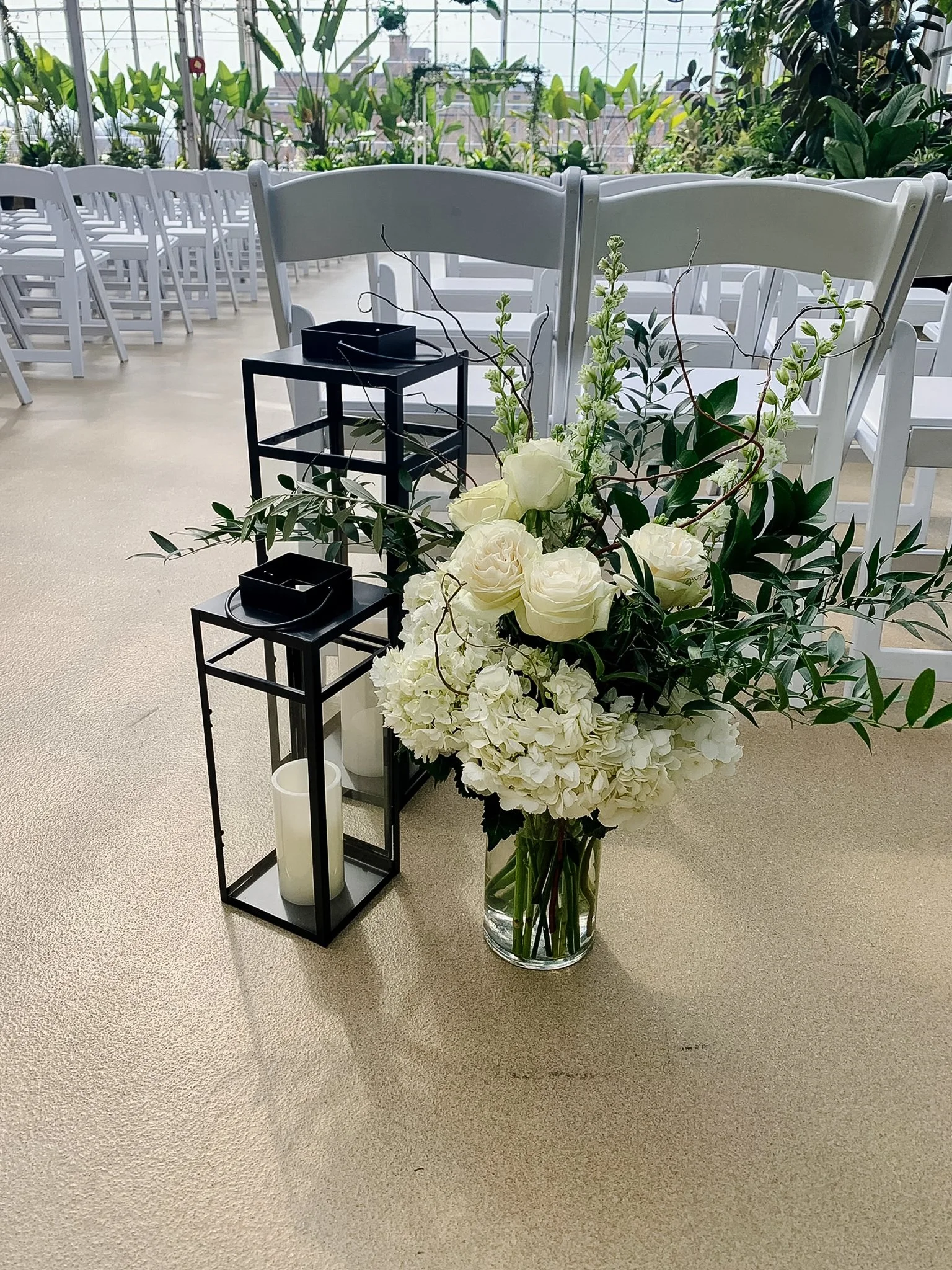 A large glass vase filled with white roses, hydrangeas, and greenery, positioned near black candle lanterns, with white chairs and plants in the background, suggesting a setup for a formal event or wedding in a greenhouse or conservatory.