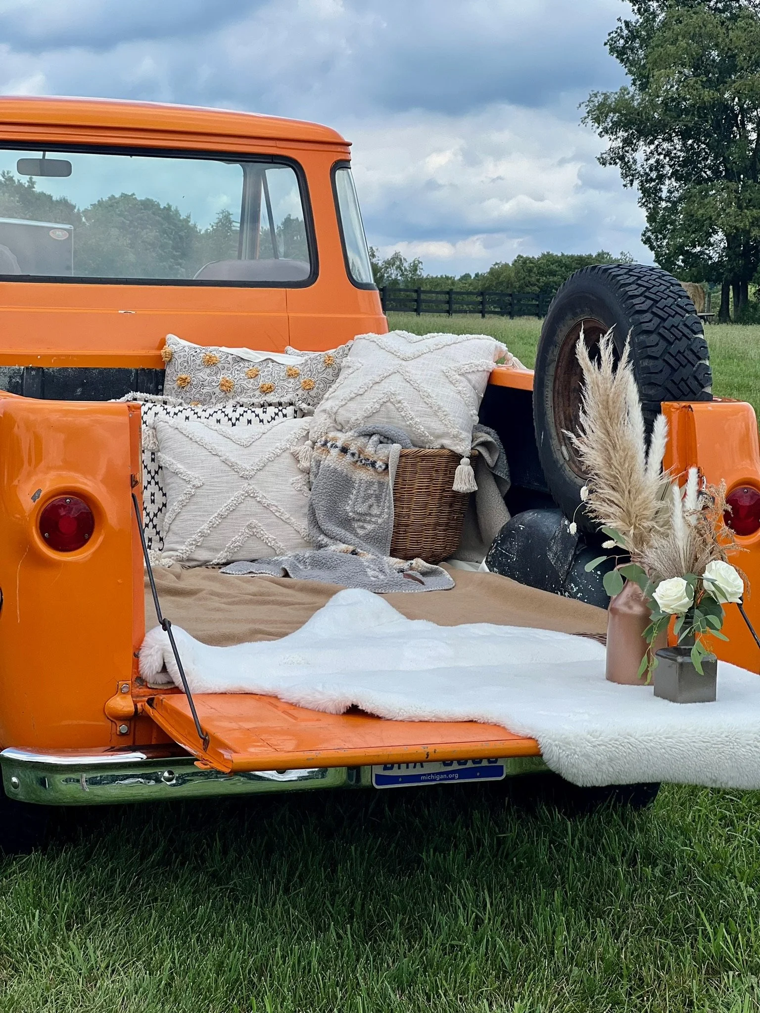 Back of an orange vintage truck with blankets, pillows, and a flower arrangement in a pot, on a grassy field with cloudy sky.
