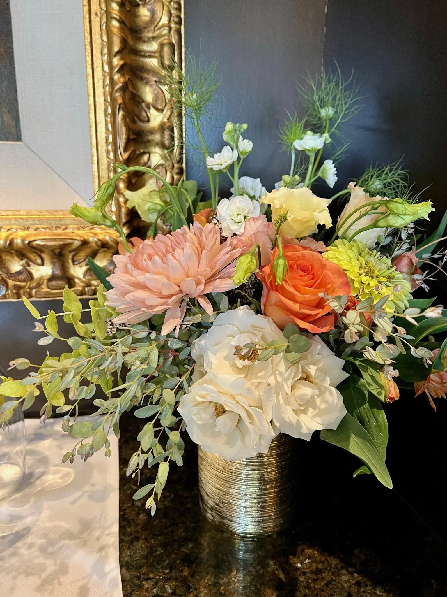 A floral arrangement in a tall, gold, textured vase placed on a dark countertop. The bouquet includes white, orange, and pink flowers, greenery, and delicate light green sprigs.