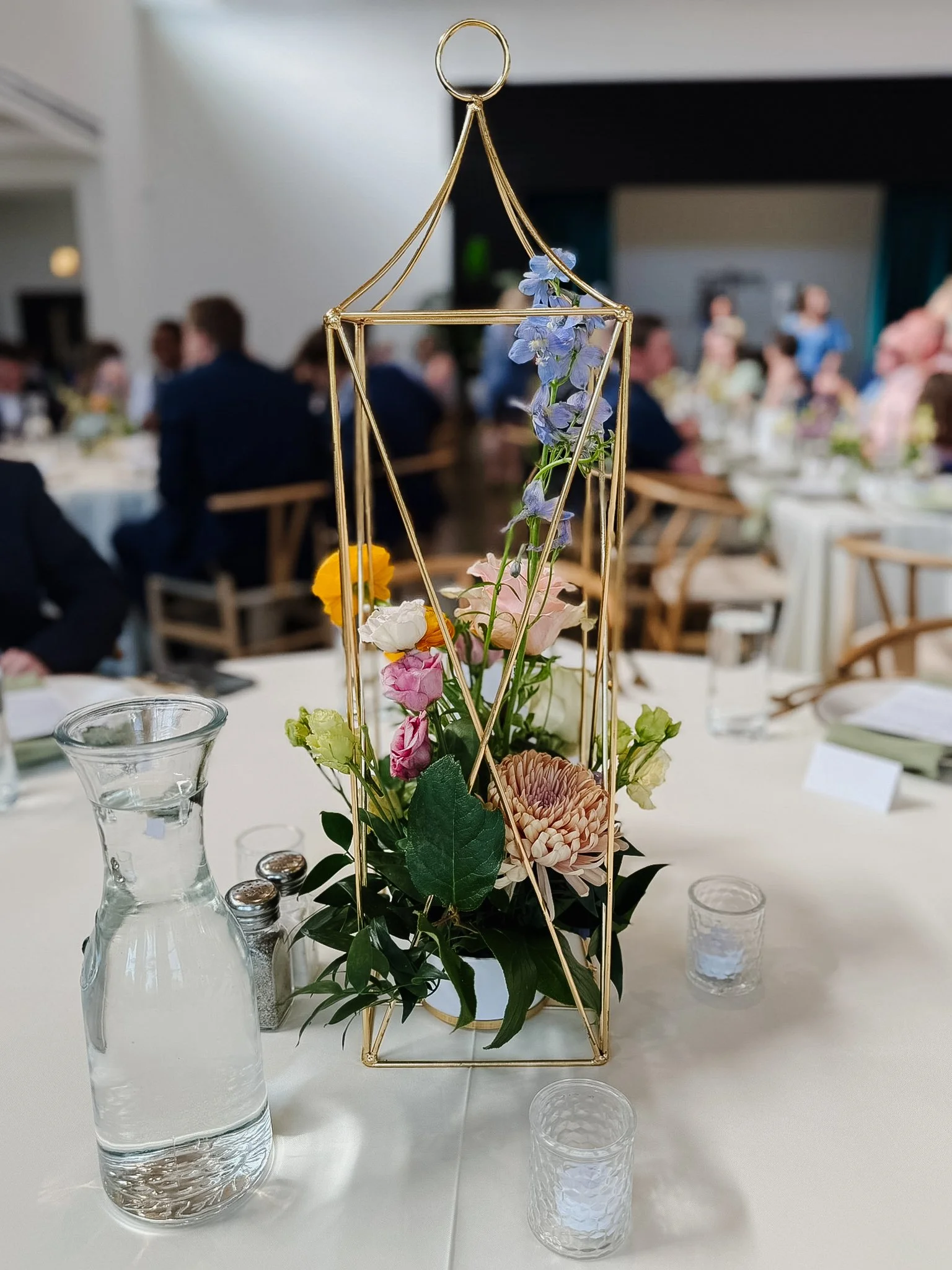 A floral centerpiece featuring blue, pink, yellow, and beige flowers inside a gold geometric vase on a white table. There are glasses and salt and pepper shakers nearby in a banquet hall with many guests seated in the background.