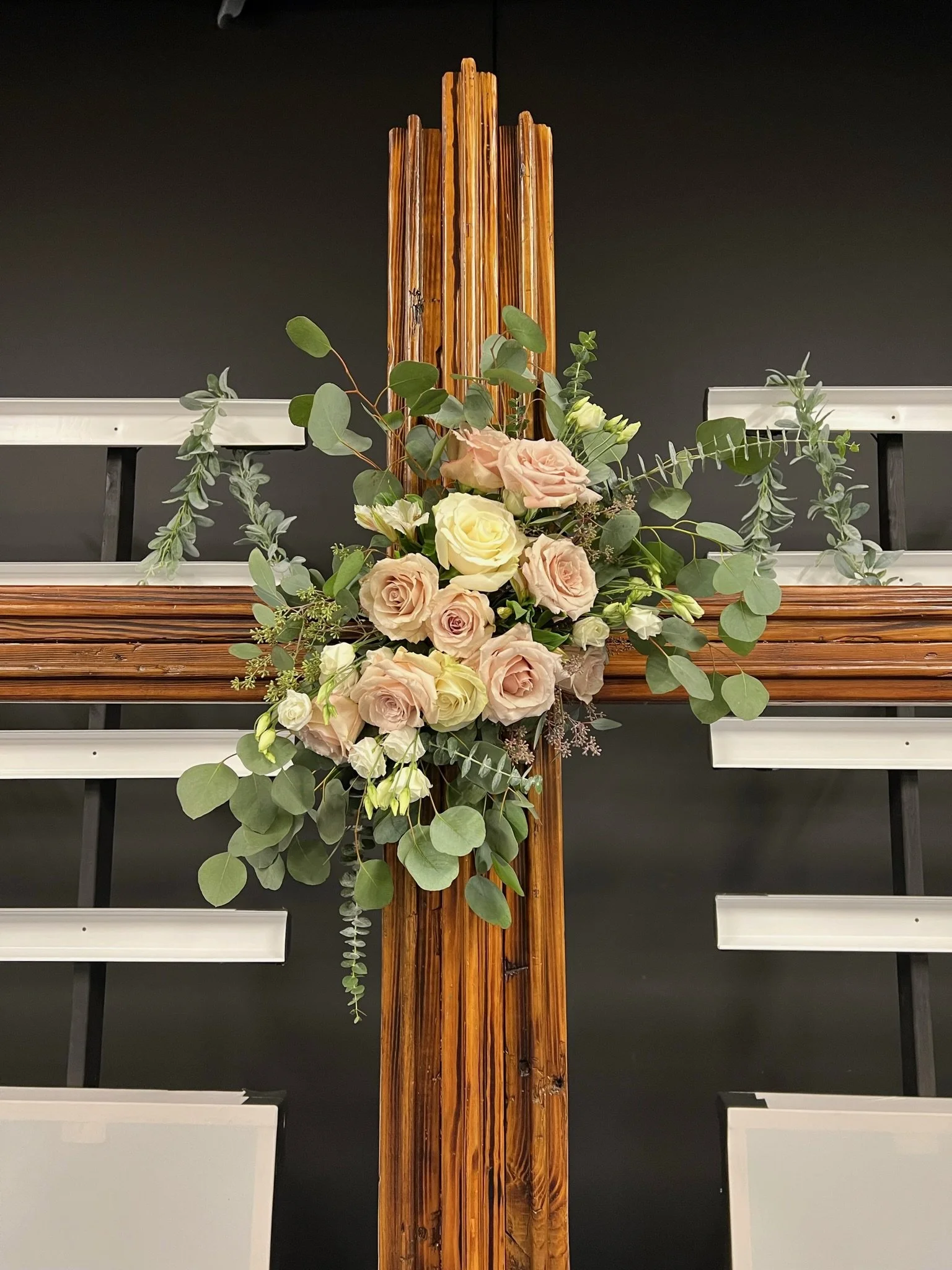 A wooden cross adorned with a floral arrangement of roses and eucalyptus, against a dark background.