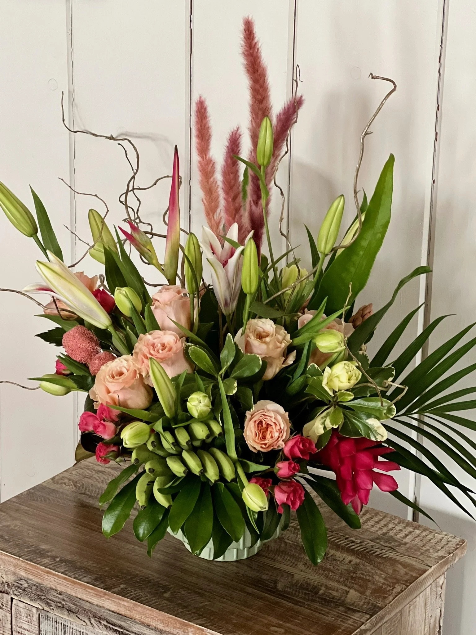 A large floral arrangement in a green striped vase on a wooden table against a white paneled wall.