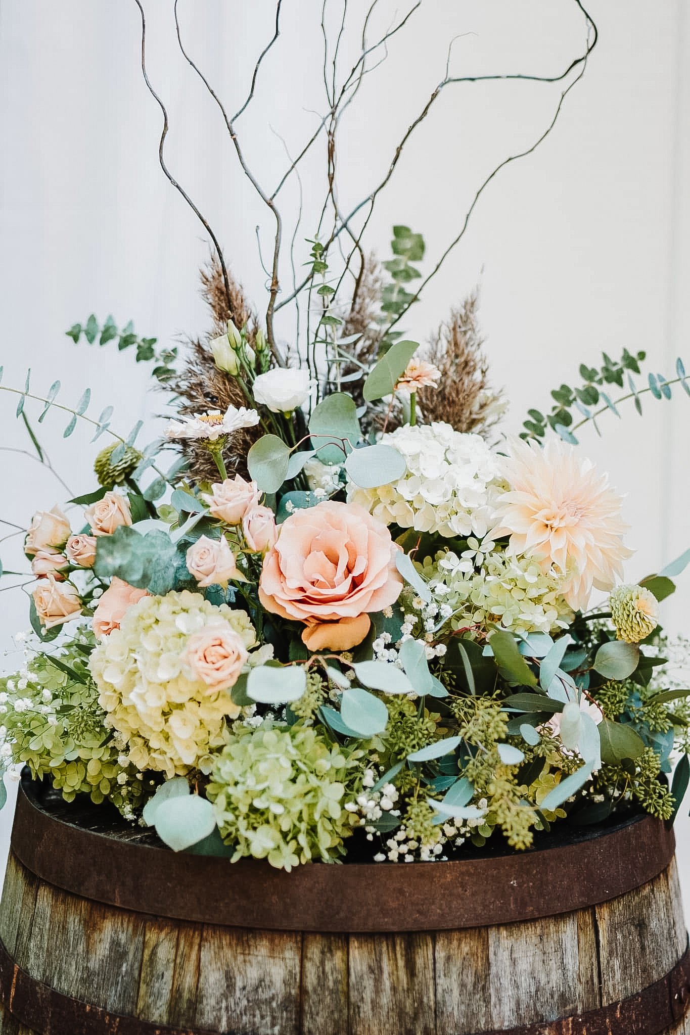 A floral arrangement featuring roses, dahlias, hydrangeas, eucalyptus, and decorative twigs in a wooden container.