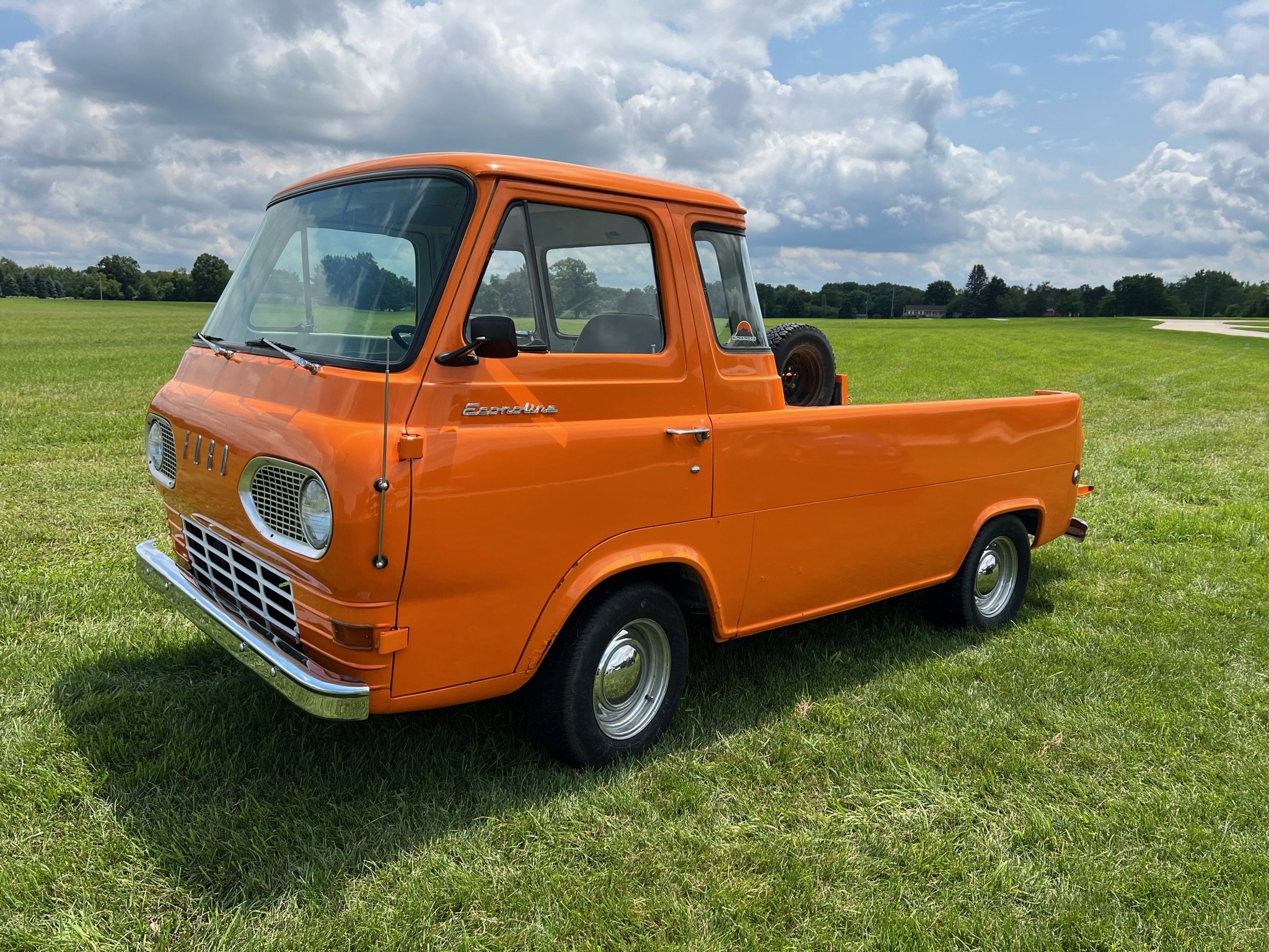 Orange vintage pickup truck parked on a grassy field with a cloudy sky in the background.