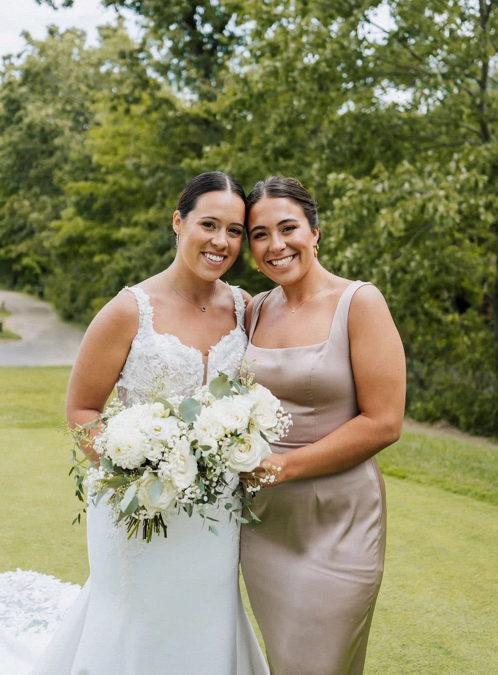Two women smiling, one in a white wedding dress holding a bouquet, and the other in a beige dress, standing outdoors with green trees in the background.
