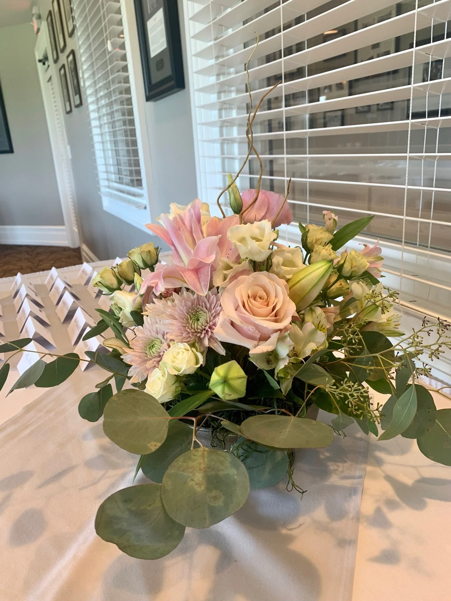 A floral arrangement with pink and white roses, pink lilies, and green foliage, placed on a white tablecloth near a window with white blinds.