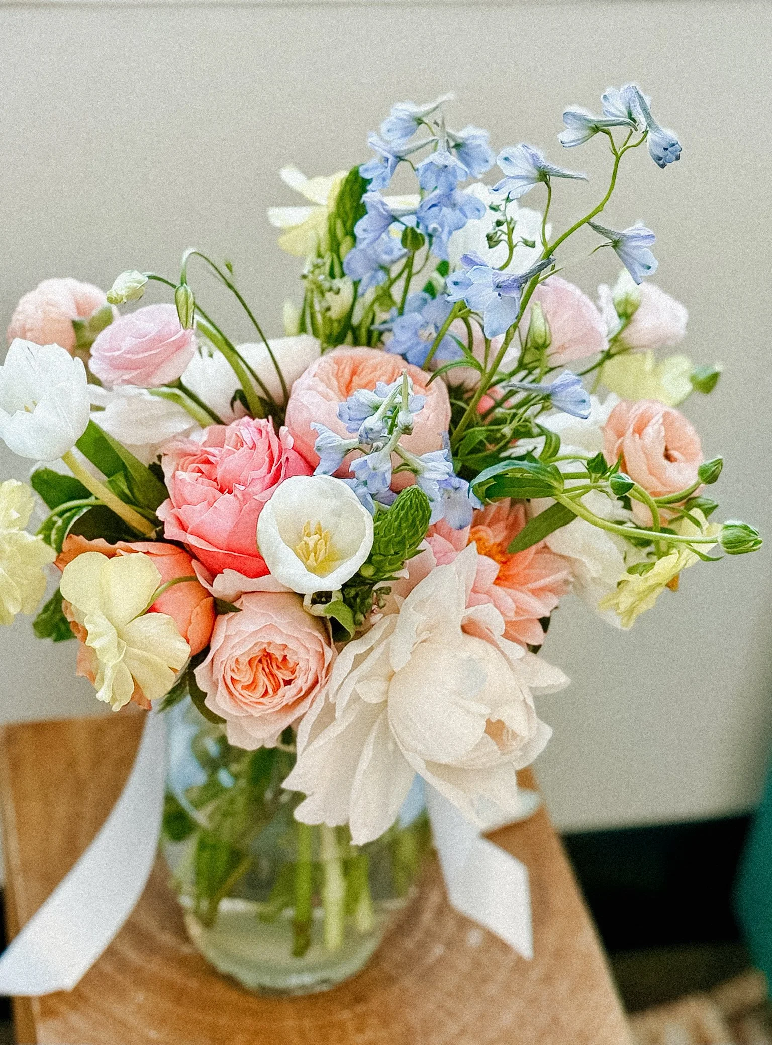 A bouquet of pastel-colored flowers, including pink, white, and light blue blooms, arranged in a glass vase with a white ribbon, held by a person's hand.