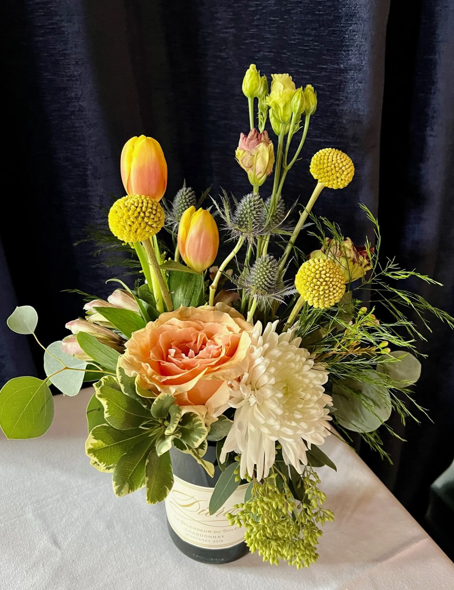 A bouquet of mixed flowers in a white paper wrapping inside a wine bottle. The bouquet includes orange tulips, yellow billy balls, green thistle, white dahlias, and greenery. The bottle has a cream and gold label.