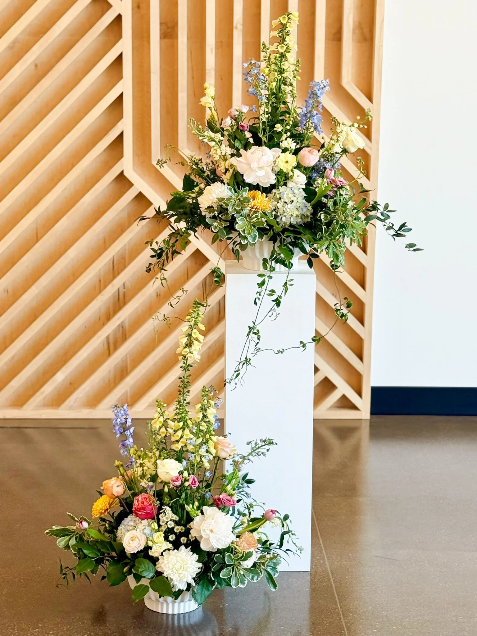 A large, colorful floral arrangement in a white striped vase, placed on a white podium, with a wooden lattice wall in the background.