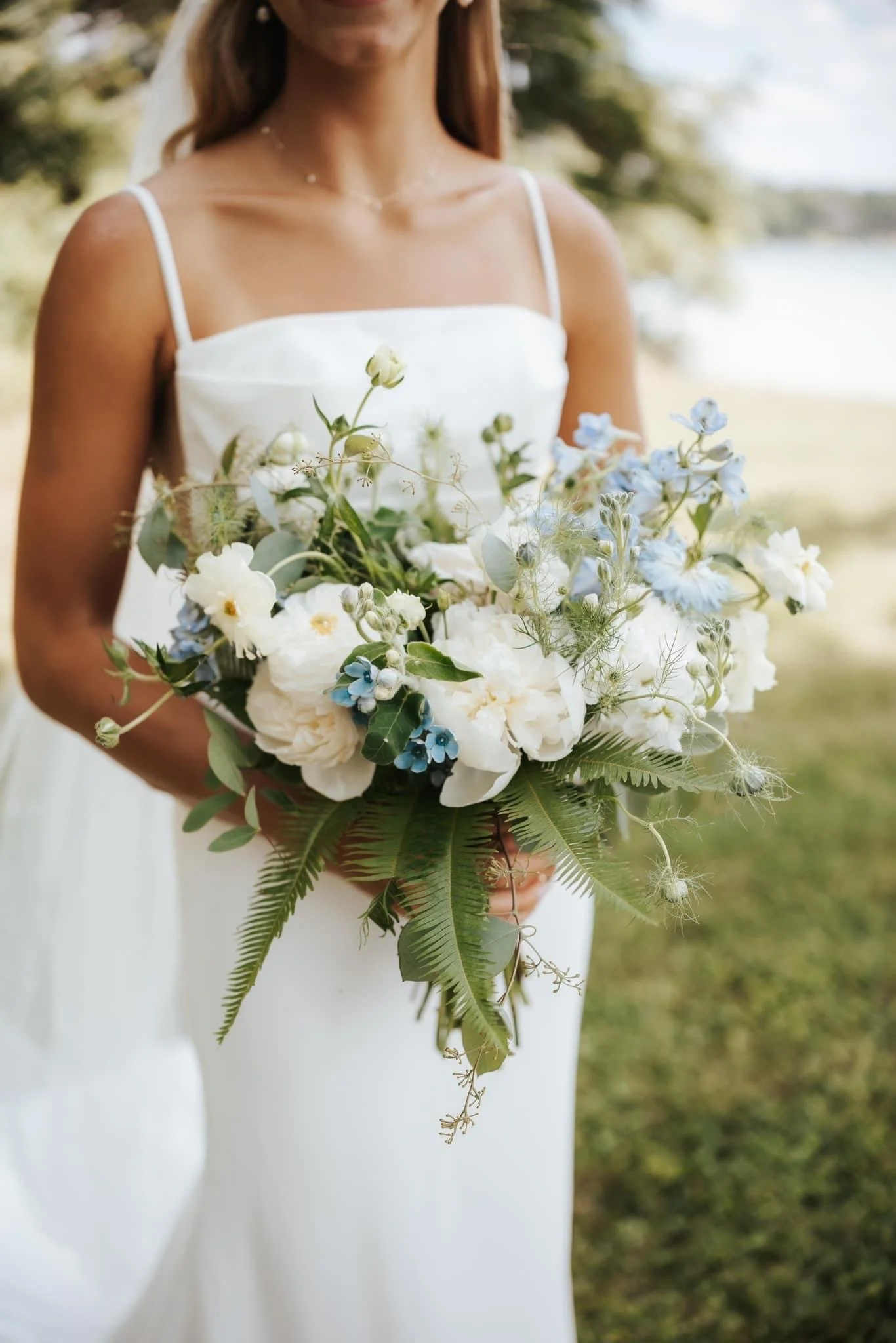 A woman in a white dress holding a bouquet of white and blue flowers outdoors with trees and water in the background.