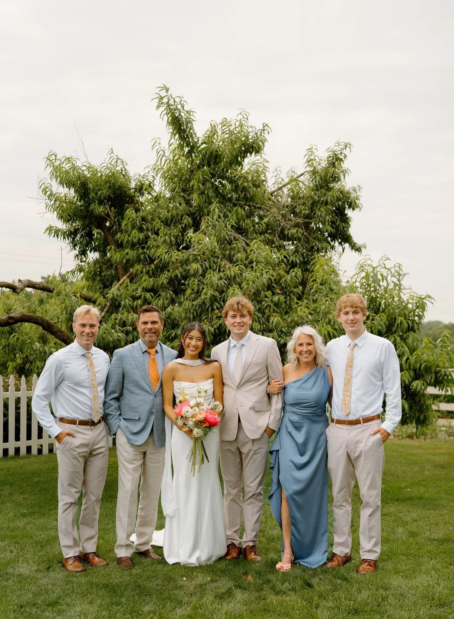 Group of seven people smiling outdoors during a wedding celebration, standing on green lawn in front of a tree with a white picket fence in the background.