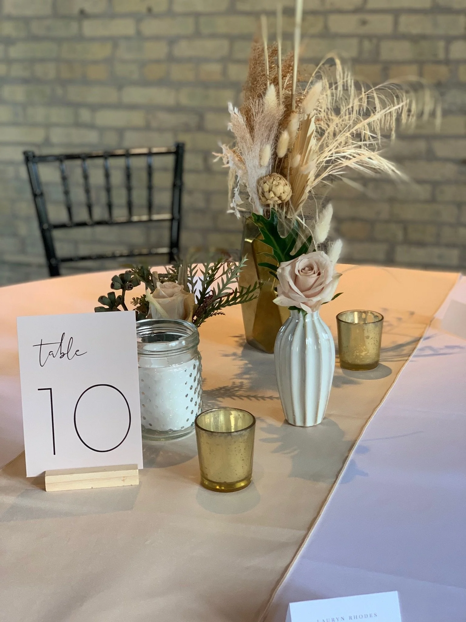 A table centerpiece with dried flowers and foliage in neutral tones, a white vase, a small candle holder, and a table number card.